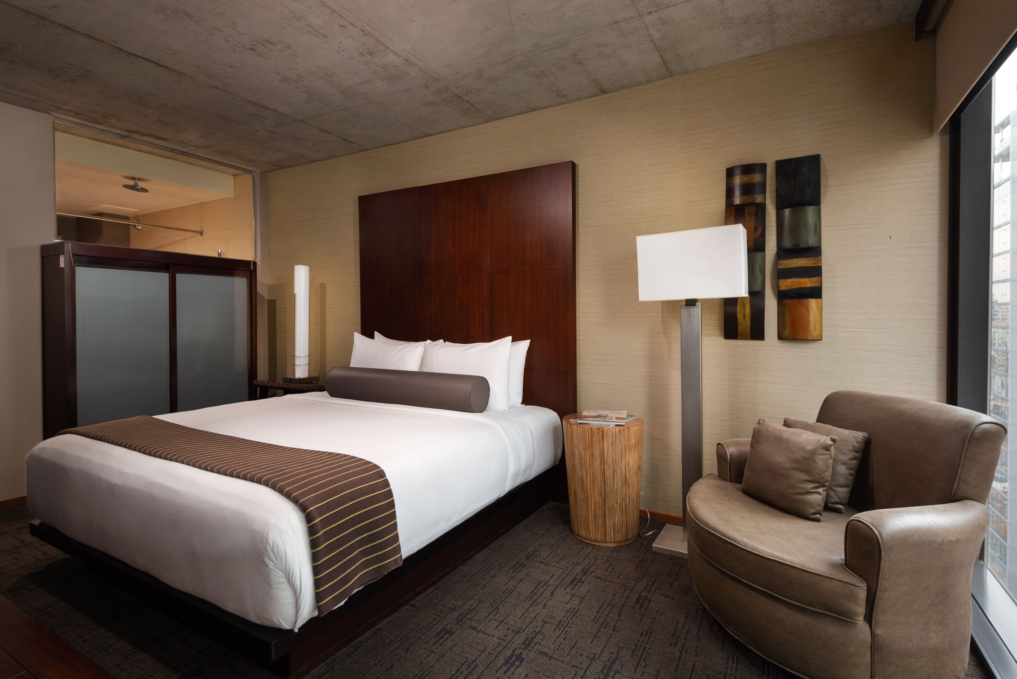 Quarto de hotel com uma cama grande, cabeceira de madeira, área de estar com uma cadeira, mesa lateral e luminária.