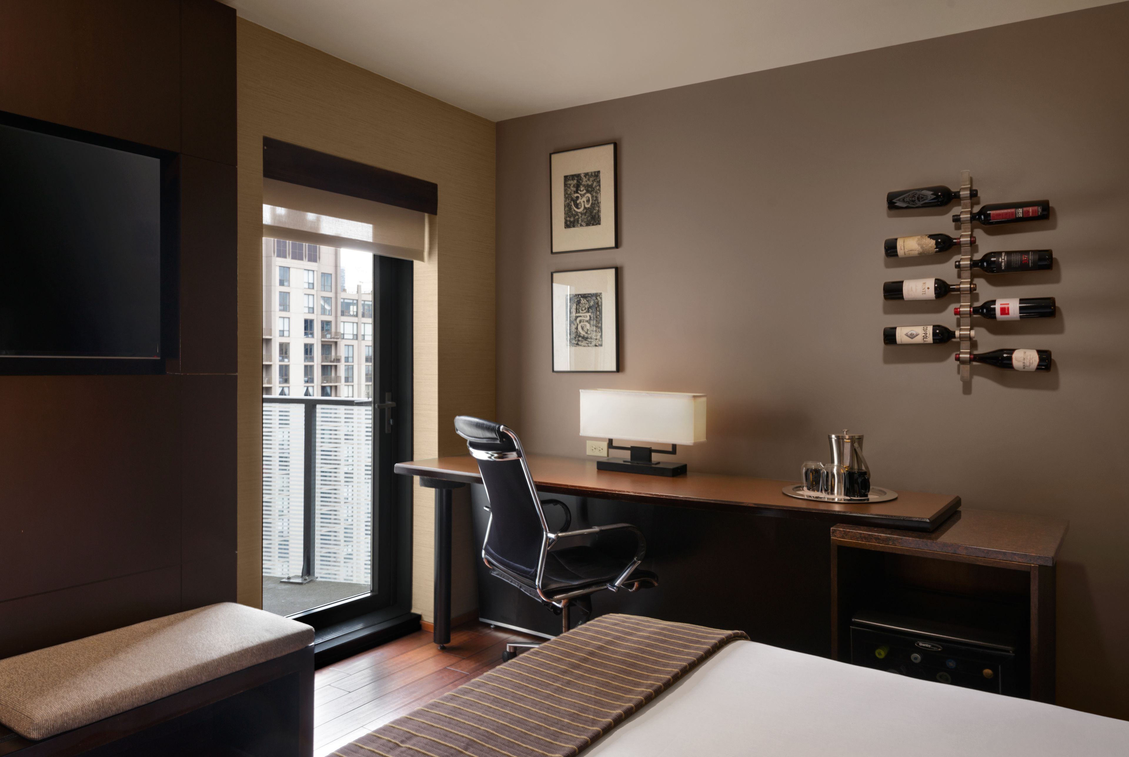 A hotel room with a bed, a desk, a chair, a TV, a window with a view of buildings, and a wall with framed pictures and wine bottles.