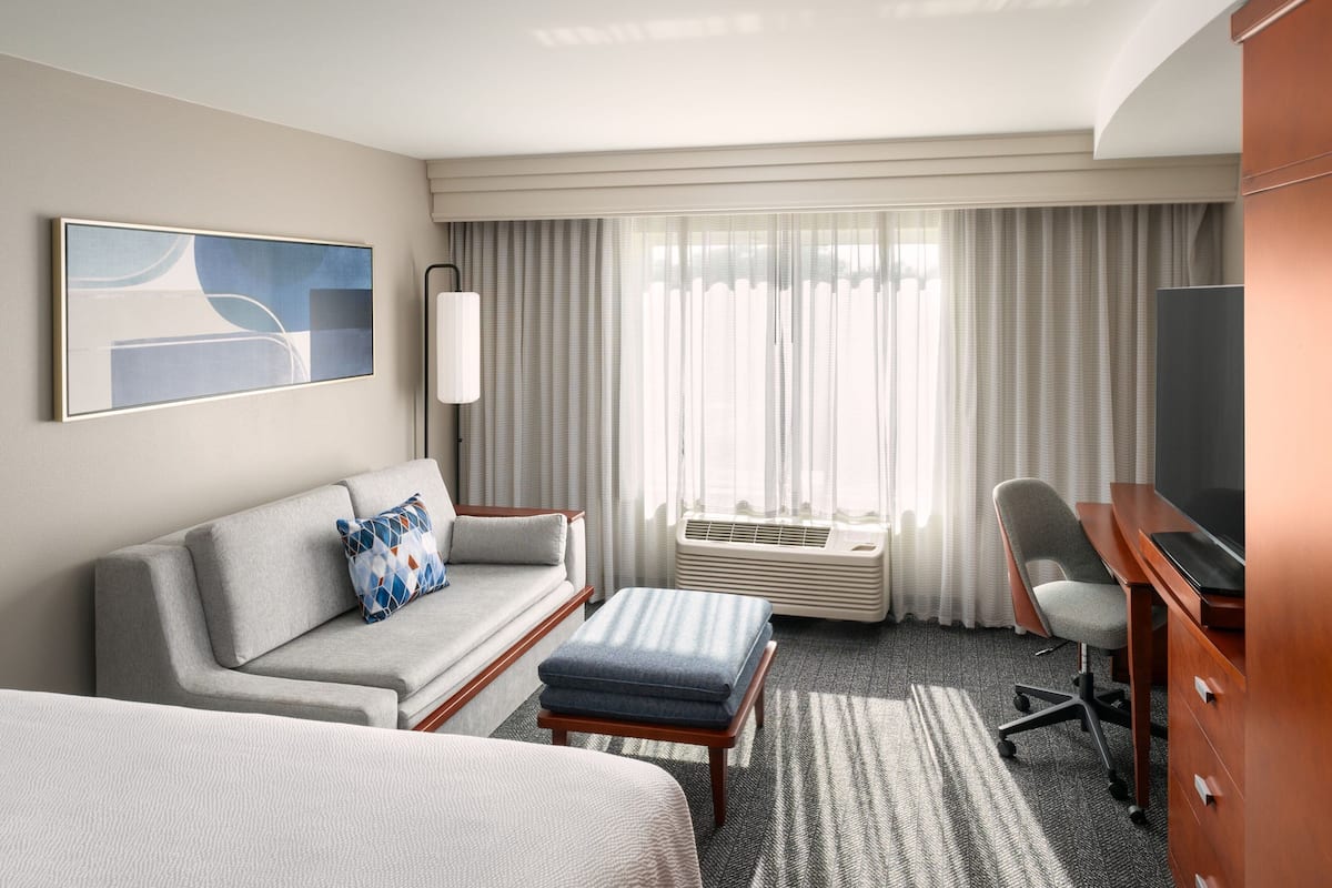 Habitación de hotel con sofá, escritorio, televisor y ventana grande con cortinas.