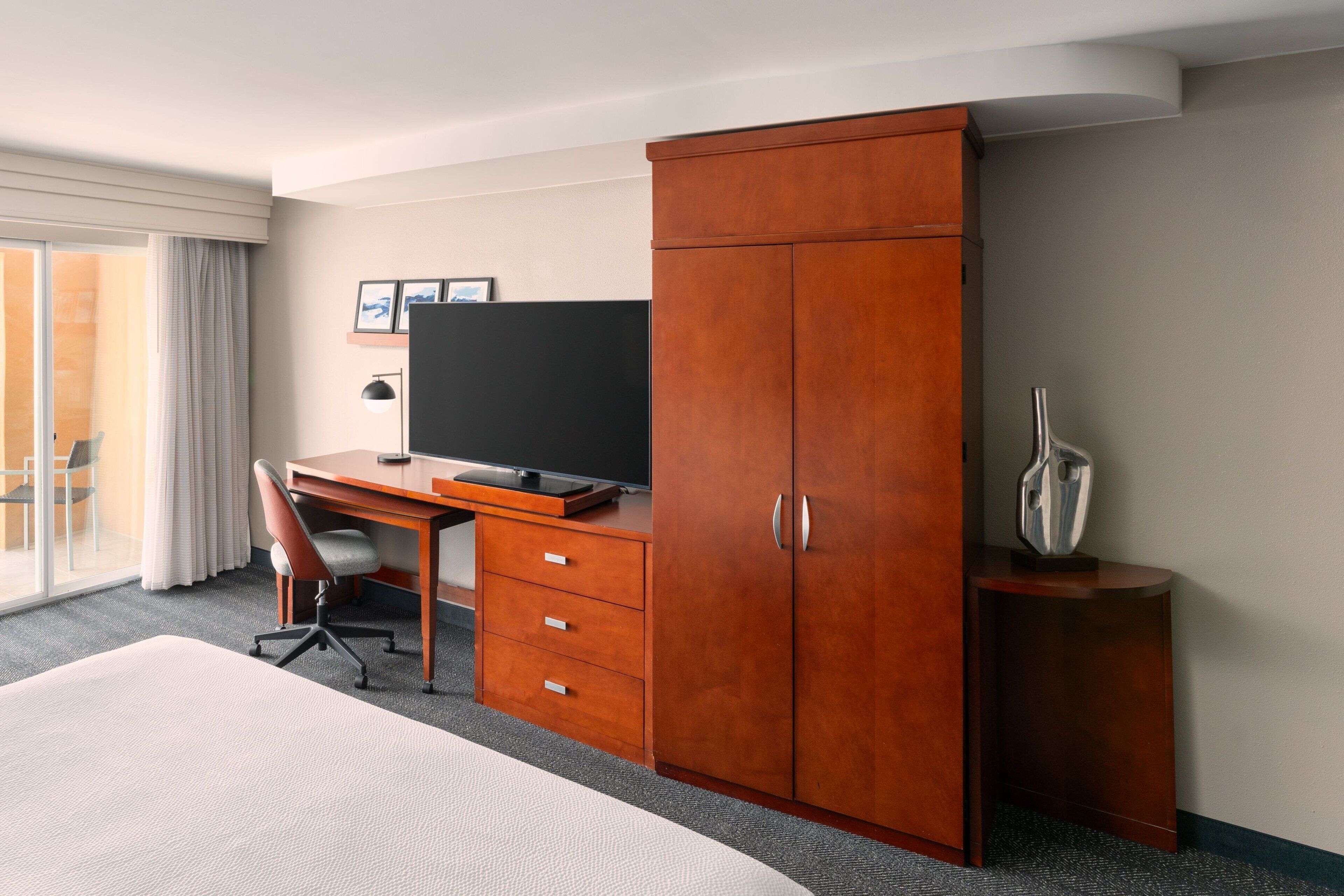 Une chambre d’hôtel comprenant un lit, un bureau avec un ordinateur, une télévision, une armoire et une petite table avec un vase.