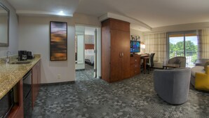 Hotelkamer met kitchenette, grote houten garderobe, televisie, grijze fauteuil en balkon met uitzicht op bomen.