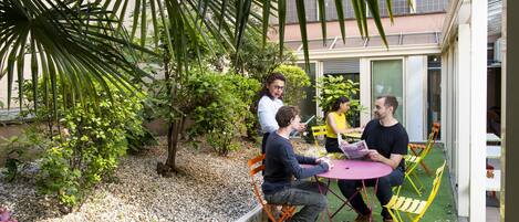 A group of people sitting at a table outdoors with a garden and a building in the background.