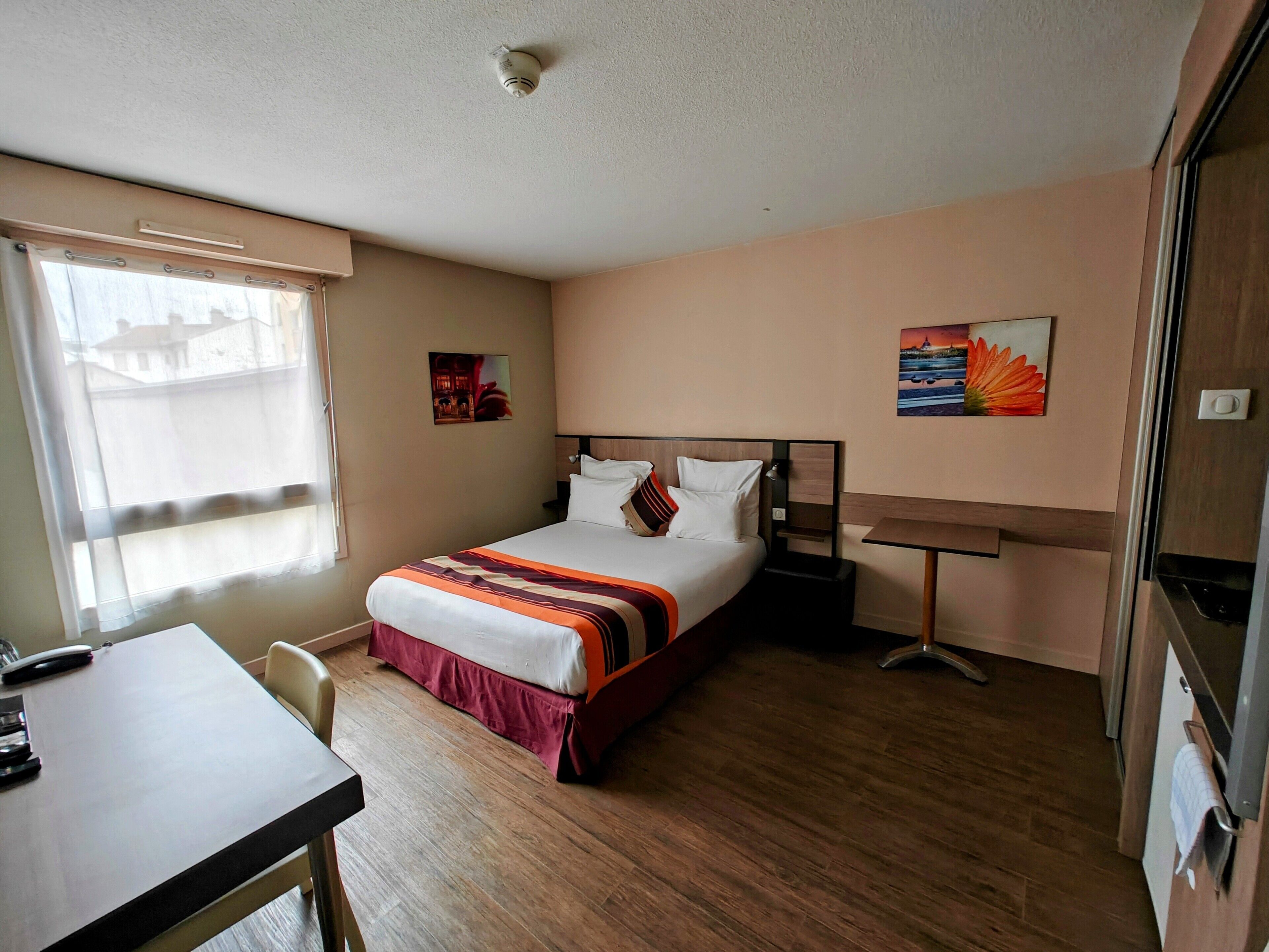 Habitación de hotel con cama, escritorio y silla. Se ve una ventana con cortinas, un cuadro en la pared y una puerta.
