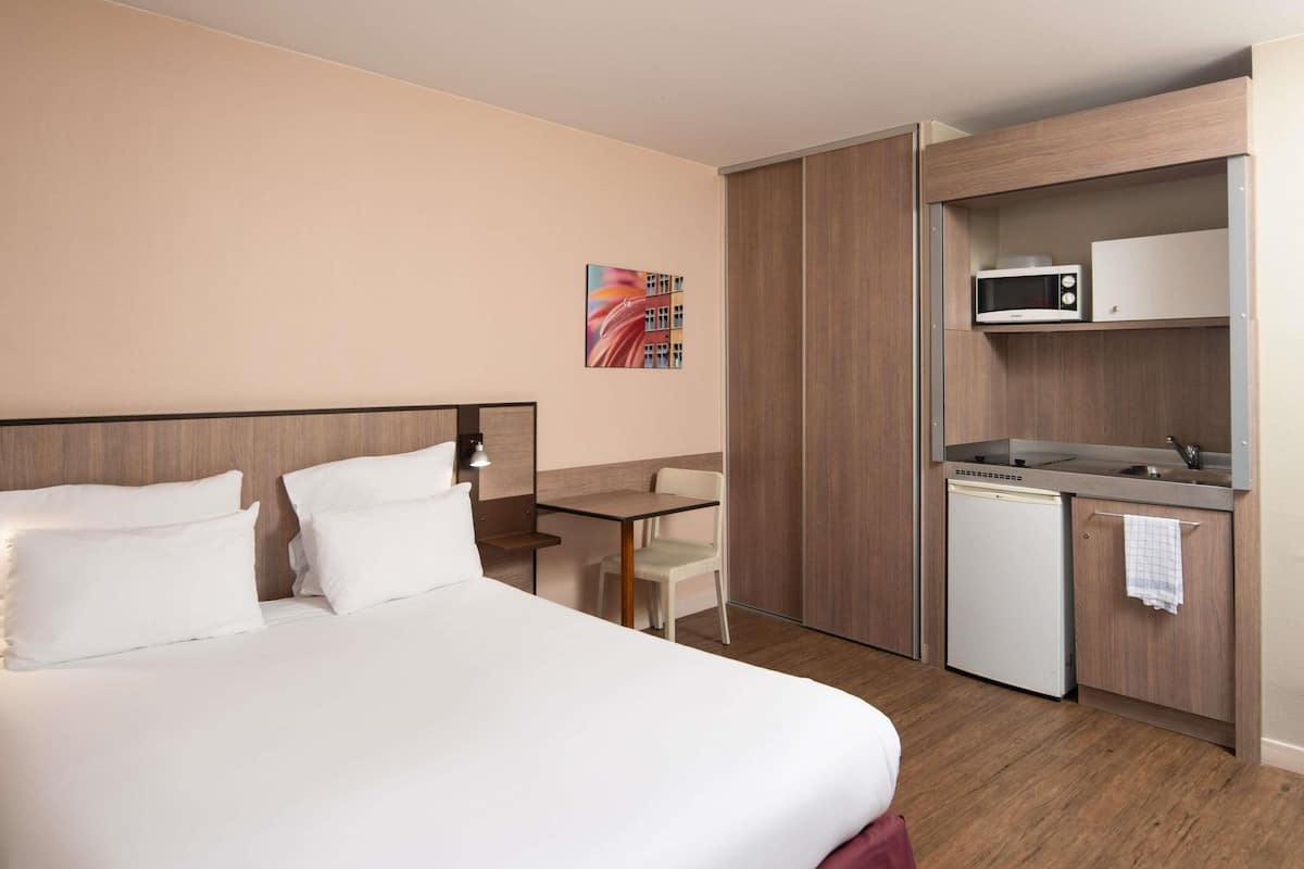 A hotel room with a bed, a desk, a small kitchenette, and a wall-mounted TV.
