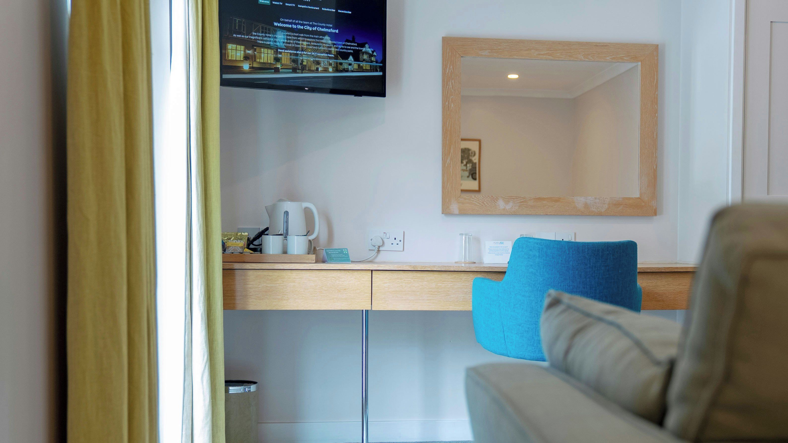 Et hotelværelse med et tv ophængt på væggen, et skrivebord med elkedel, kop og et lille skilt, en blå stol og et spejl med træramme.
