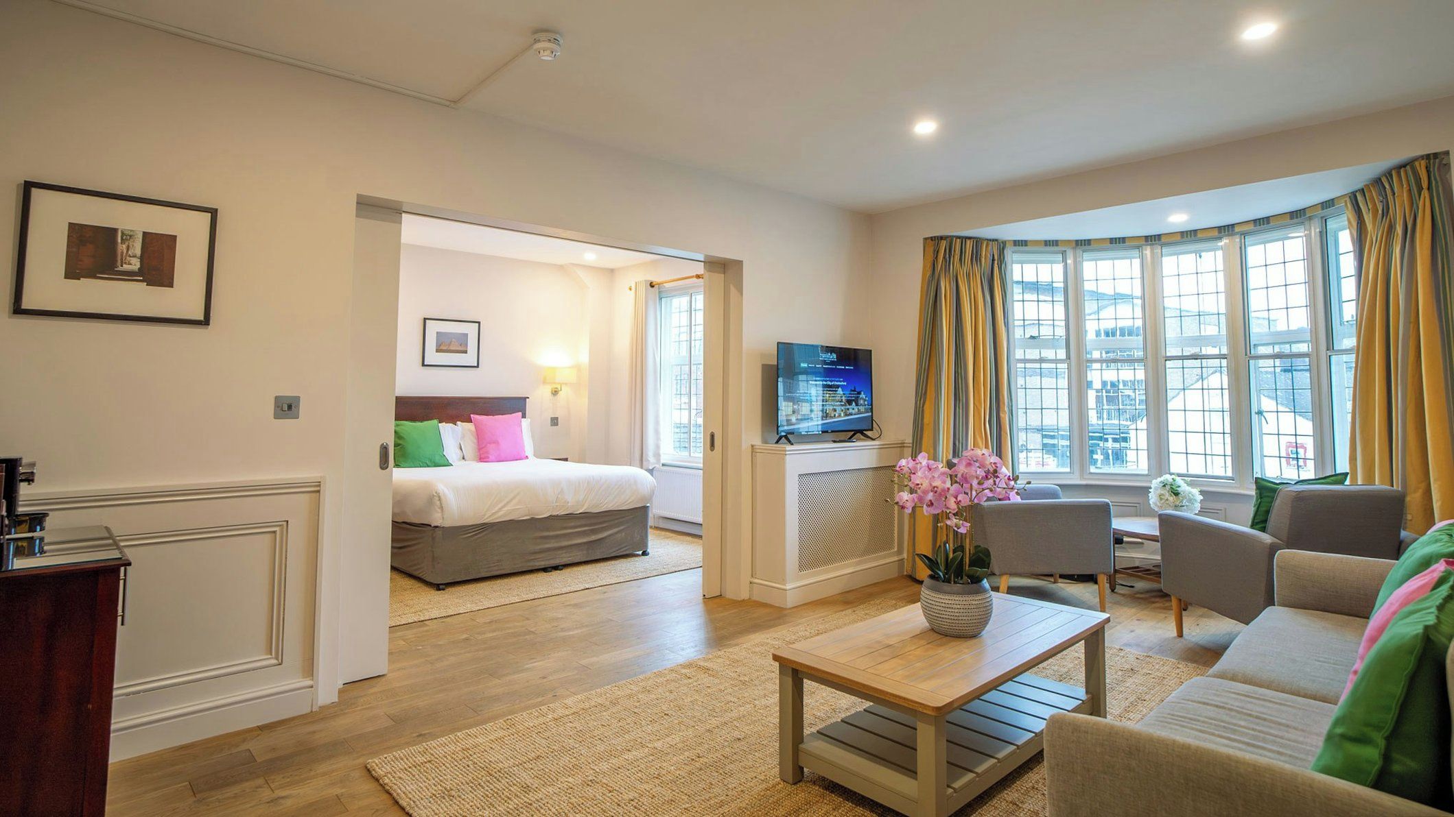 A hotel room with a bed, a TV, a sofa, a coffee table, and a view of the city through large windows.