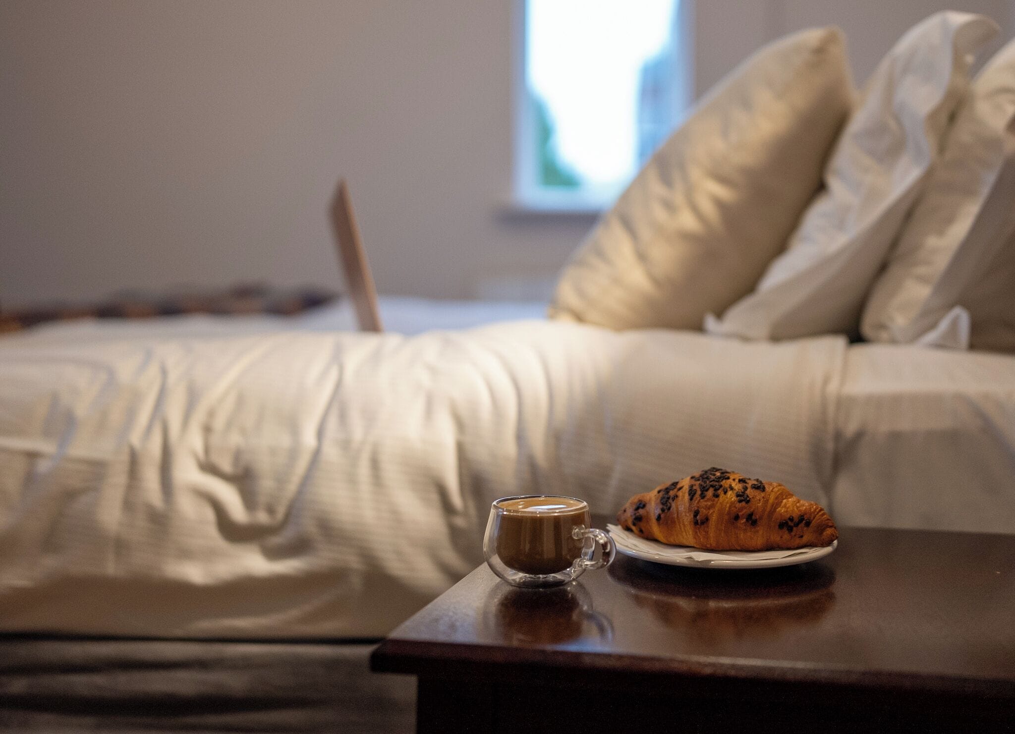 A bed with a laptop, a cup of coffee, and a croissant on a bedside table.