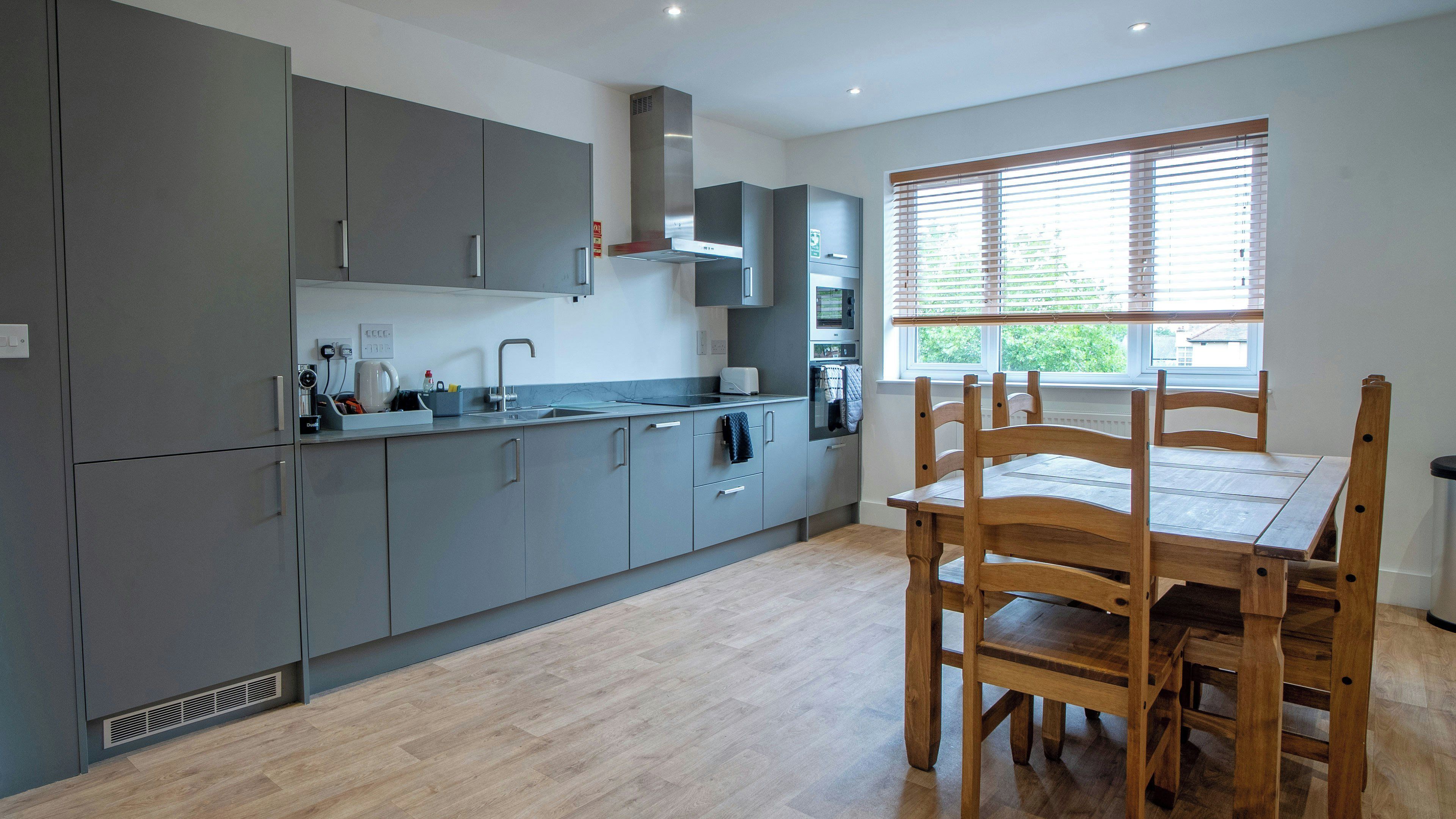 A modern kitchen with grey cabinets, a wooden dining table, and a window with blinds.