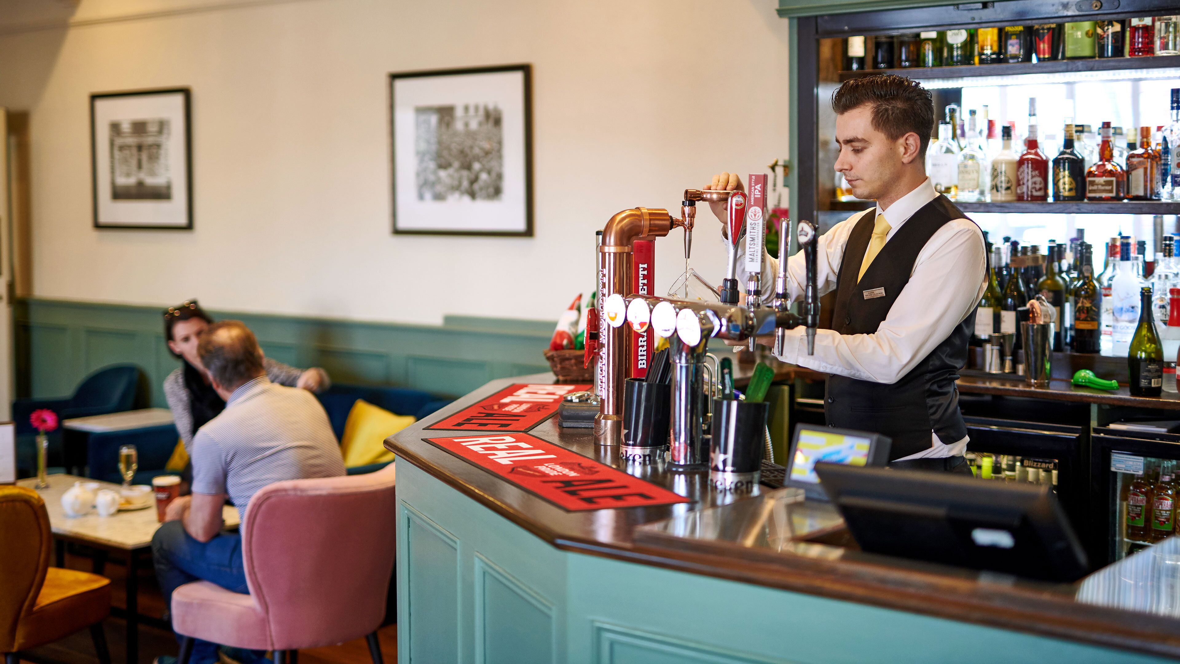 A bar area with a bartender, a counter with various bottles, and a seating area with tables and chairs.