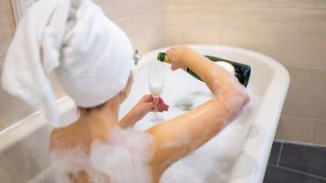 Una persona en una bañera llena de burbujas, sosteniendo una copa y sirviéndose champán.