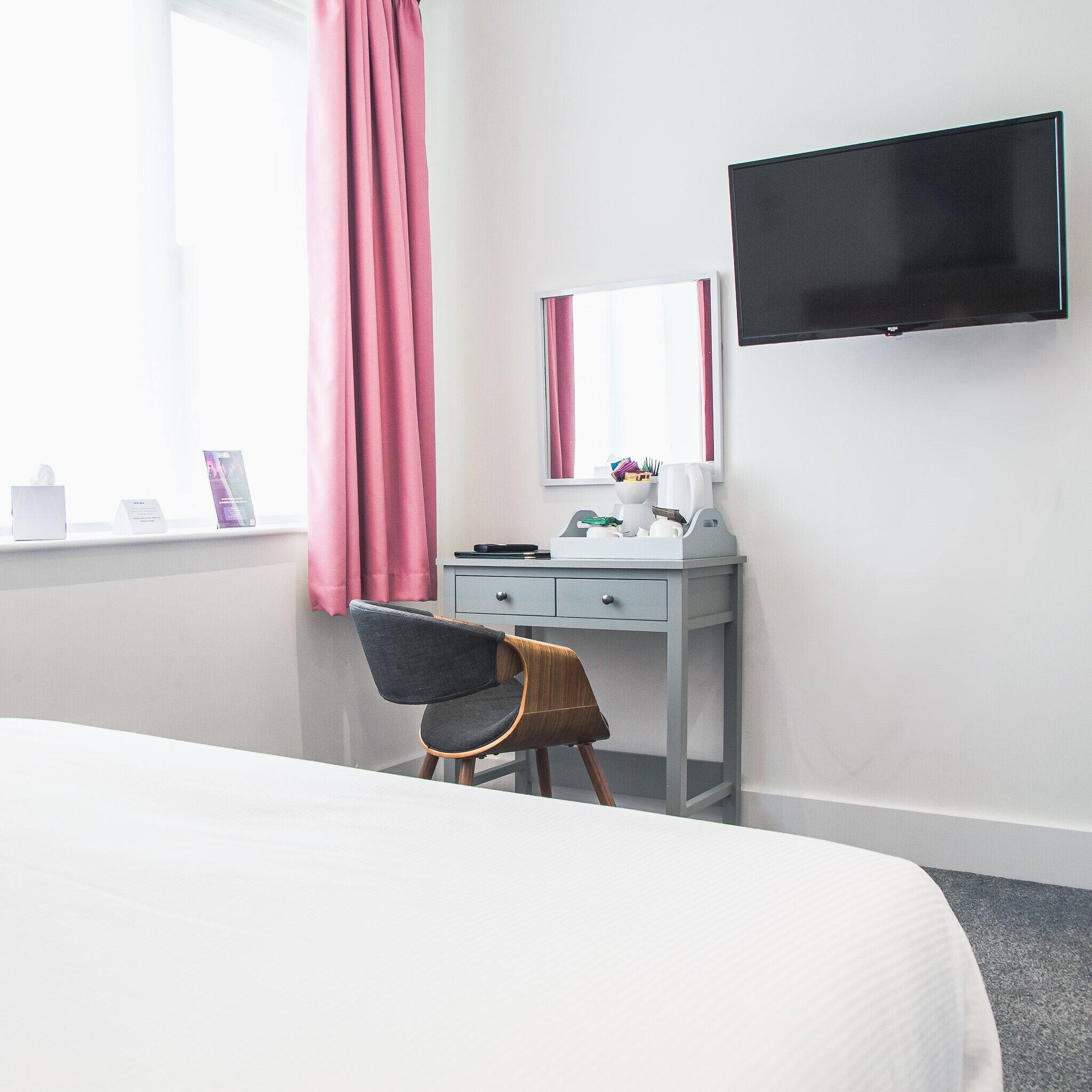 A hotel room with a bed, a desk with a chair, a flat-screen TV, and a window with curtains.