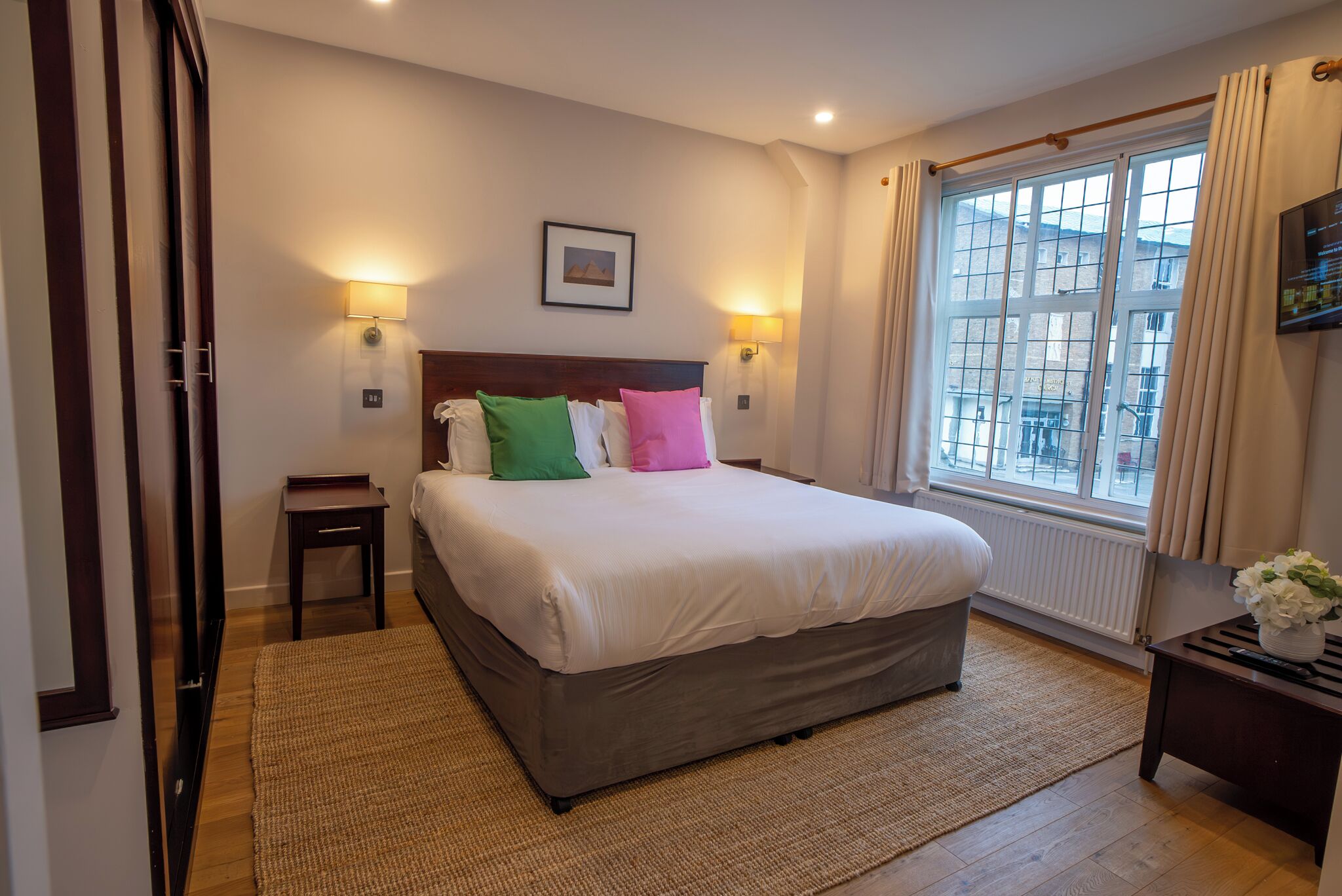 Une chambre à coucher avec un grand lit, deux tables de chevet, une commode, une fenêtre avec des rideaux et un tableau au mur.