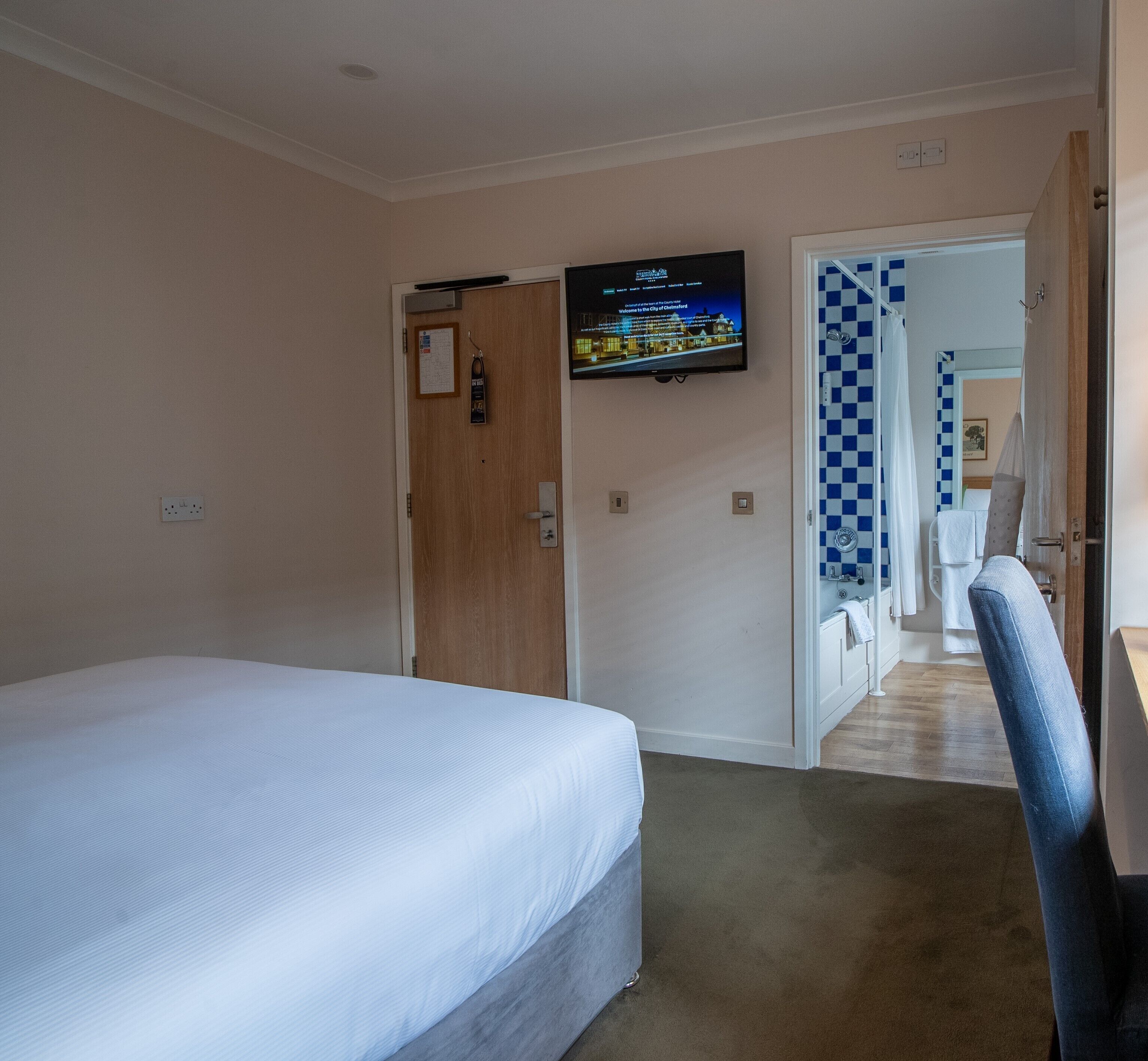 Habitación de hotel con cama, televisor en la pared, puerta que da a un baño y una silla.