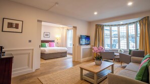 A modern living room with a sofa, coffee table, and a TV. There is a bed in the background and a window with curtains. - County Hotel Chelmsford (Chelmsford)