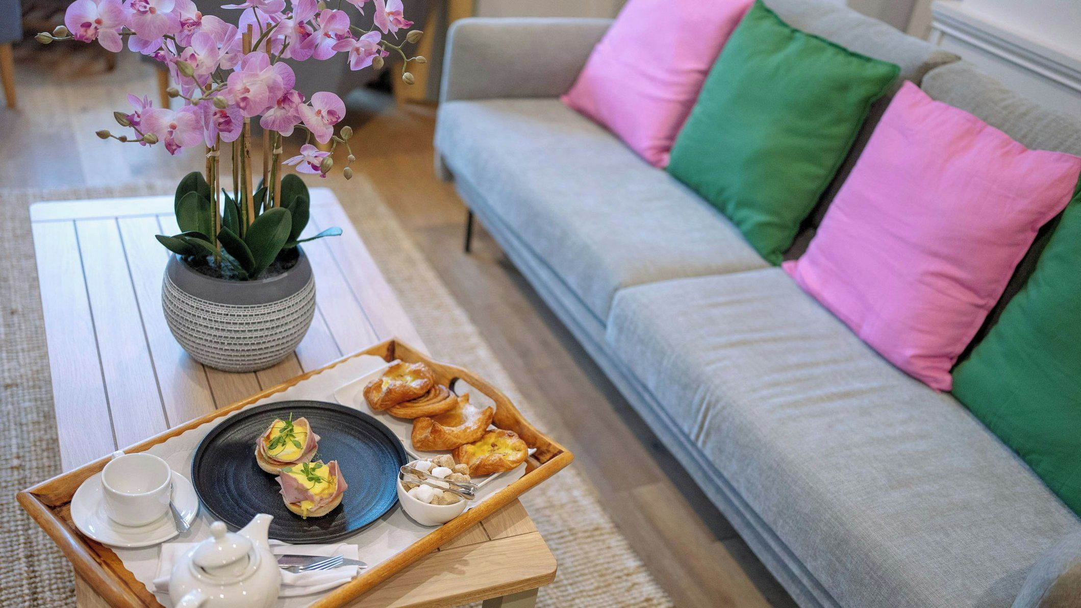 A tray with food and drinks on a wooden table, a sofa with colorful cushions, and a potted plant with flowers.