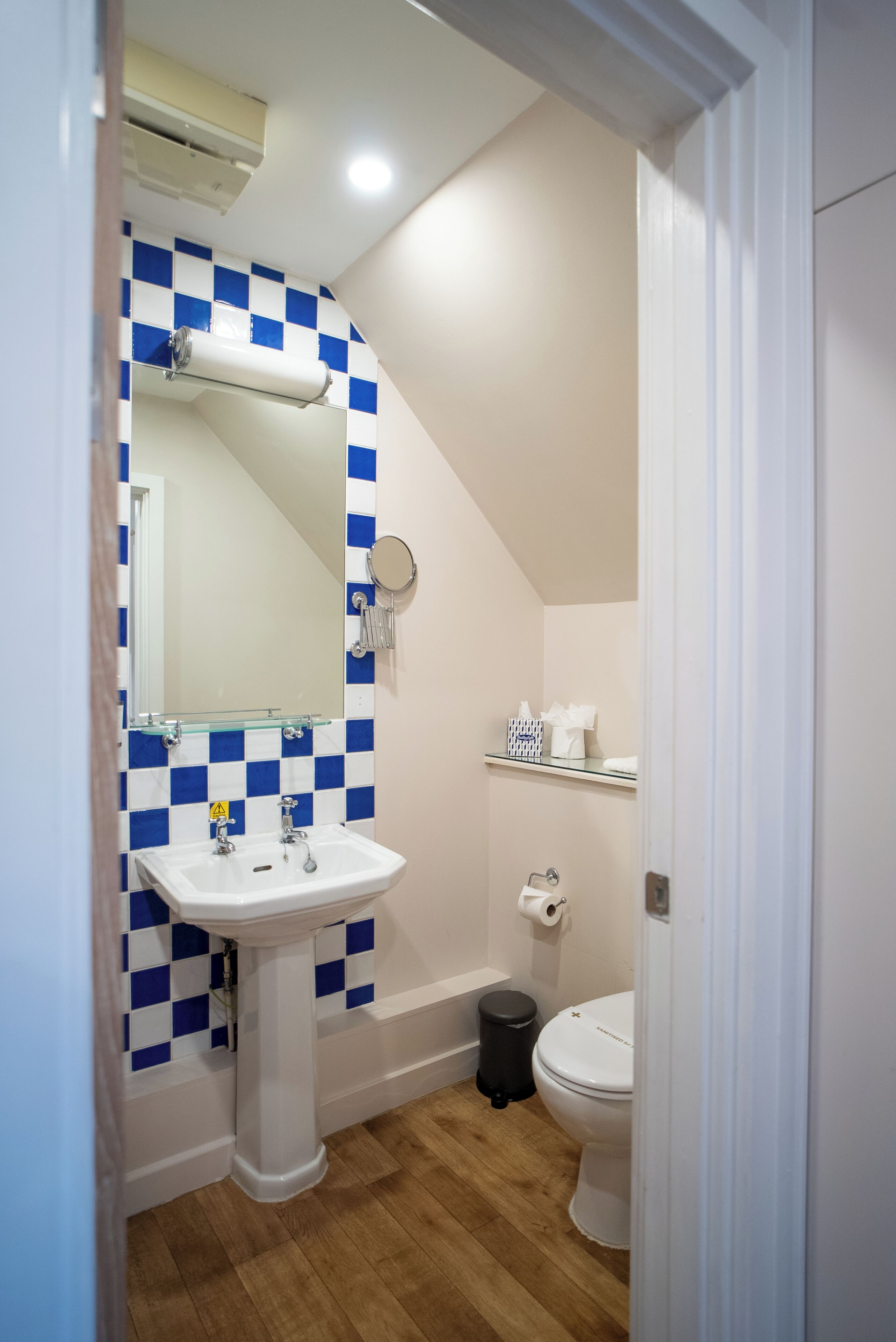 Et badeværelse med et skakmønstret backsplash i blå og hvide fliser, en hvid konsolvask, et spejl, et toilet og en sort skraldespand.