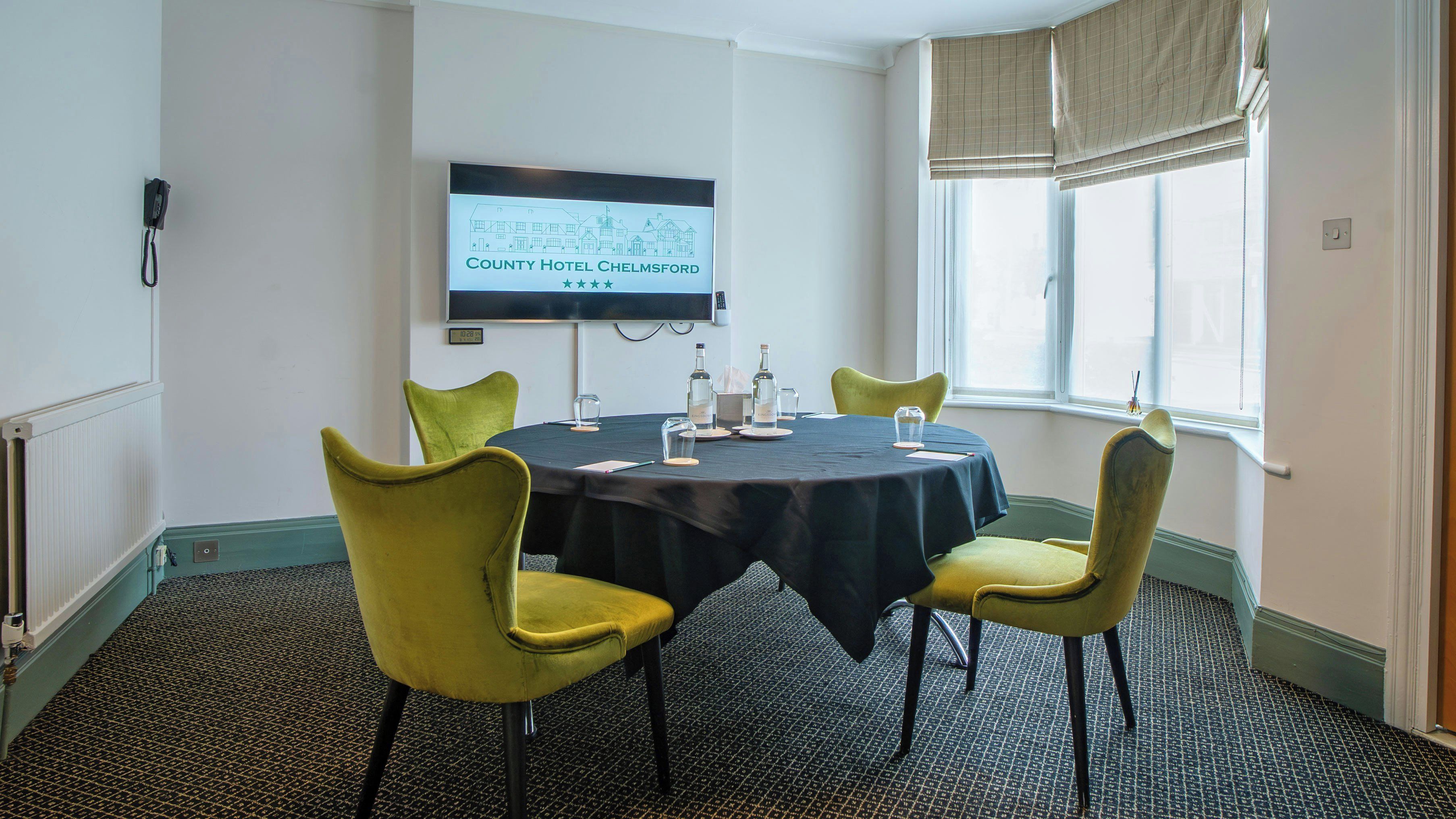 A conference room with a black table, yellow chairs, a television screen, and a window with blinds.