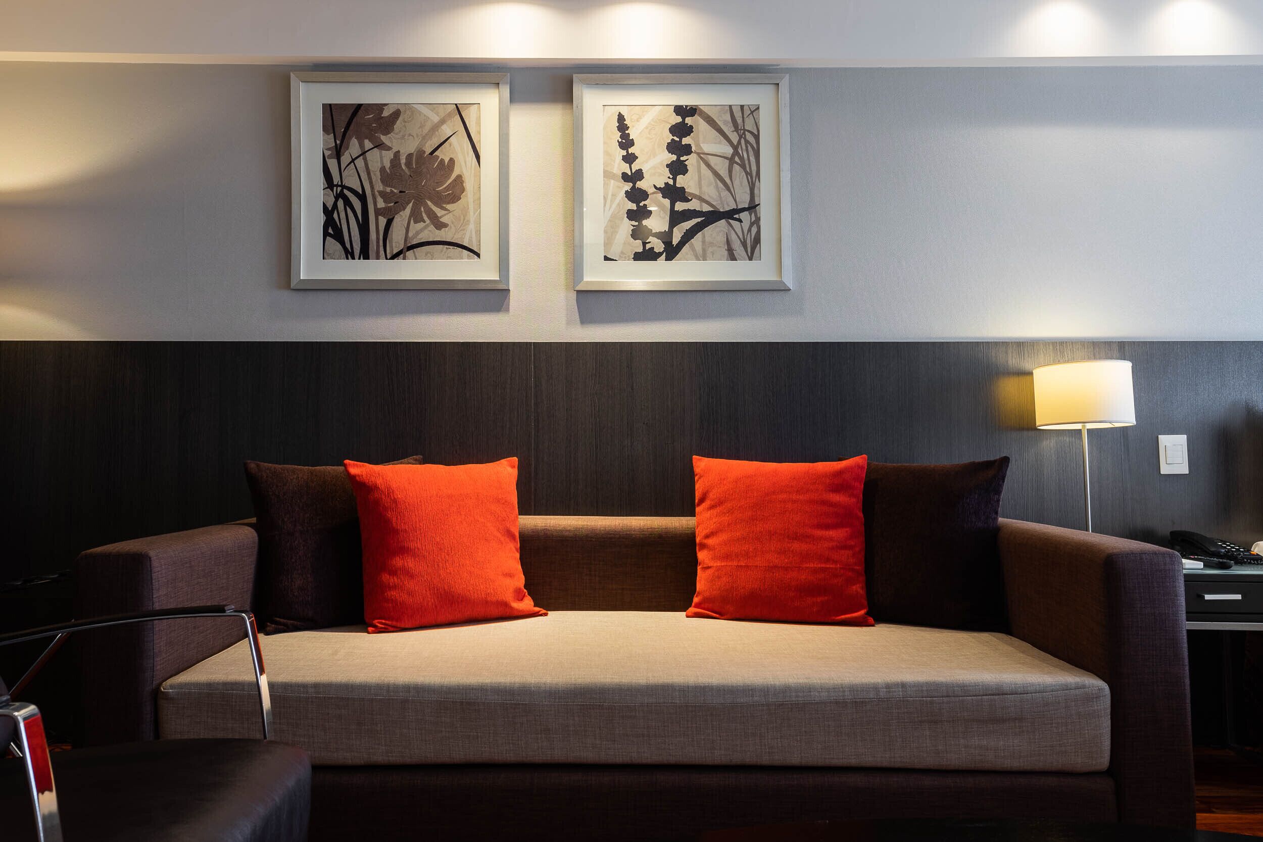 A modern hotel room with a sofa, two framed floral artworks, and a bedside lamp.
