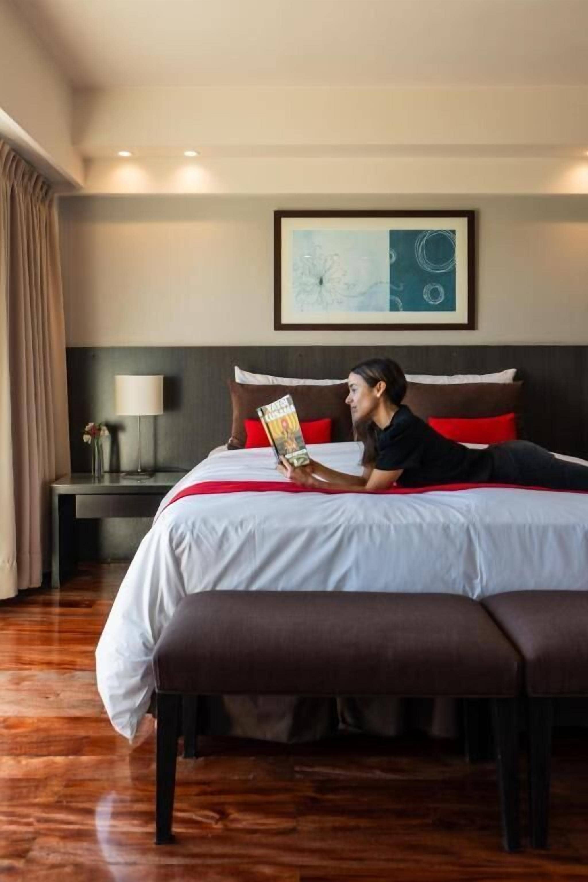 Una habitación de hotel con una cama, una mesita de noche con lámpara, una obra de arte enmarcada en la pared y un ventanal con cortinas.