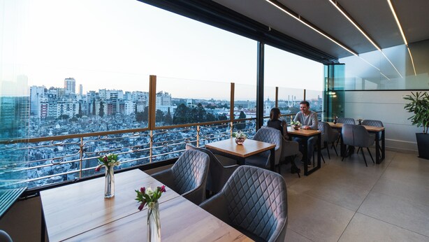 Een moderne eetruimte met grote ramen en uitzicht over de stad, tafels met stoelen, en een vaas met bloemen op tafel.