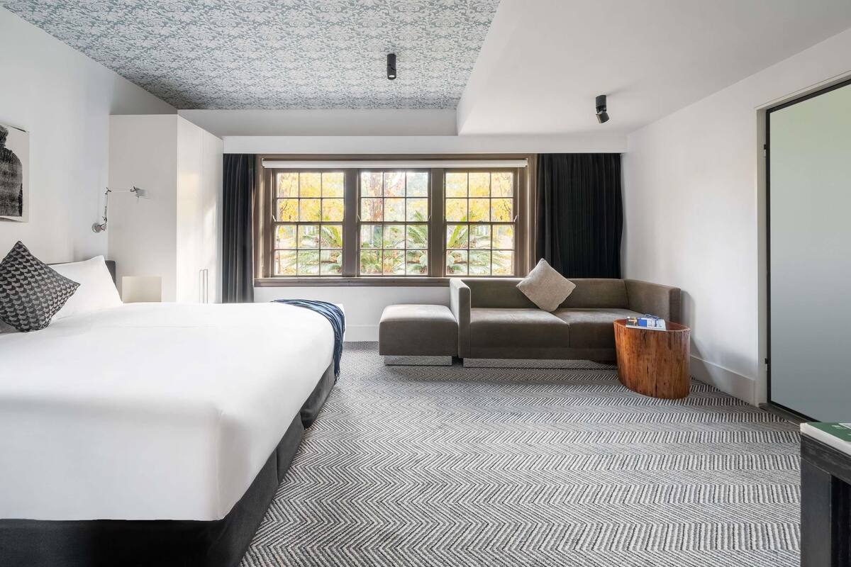 A hotel room with a bed, a sofa, a window with a view of trees, and a ceiling with a floral pattern.