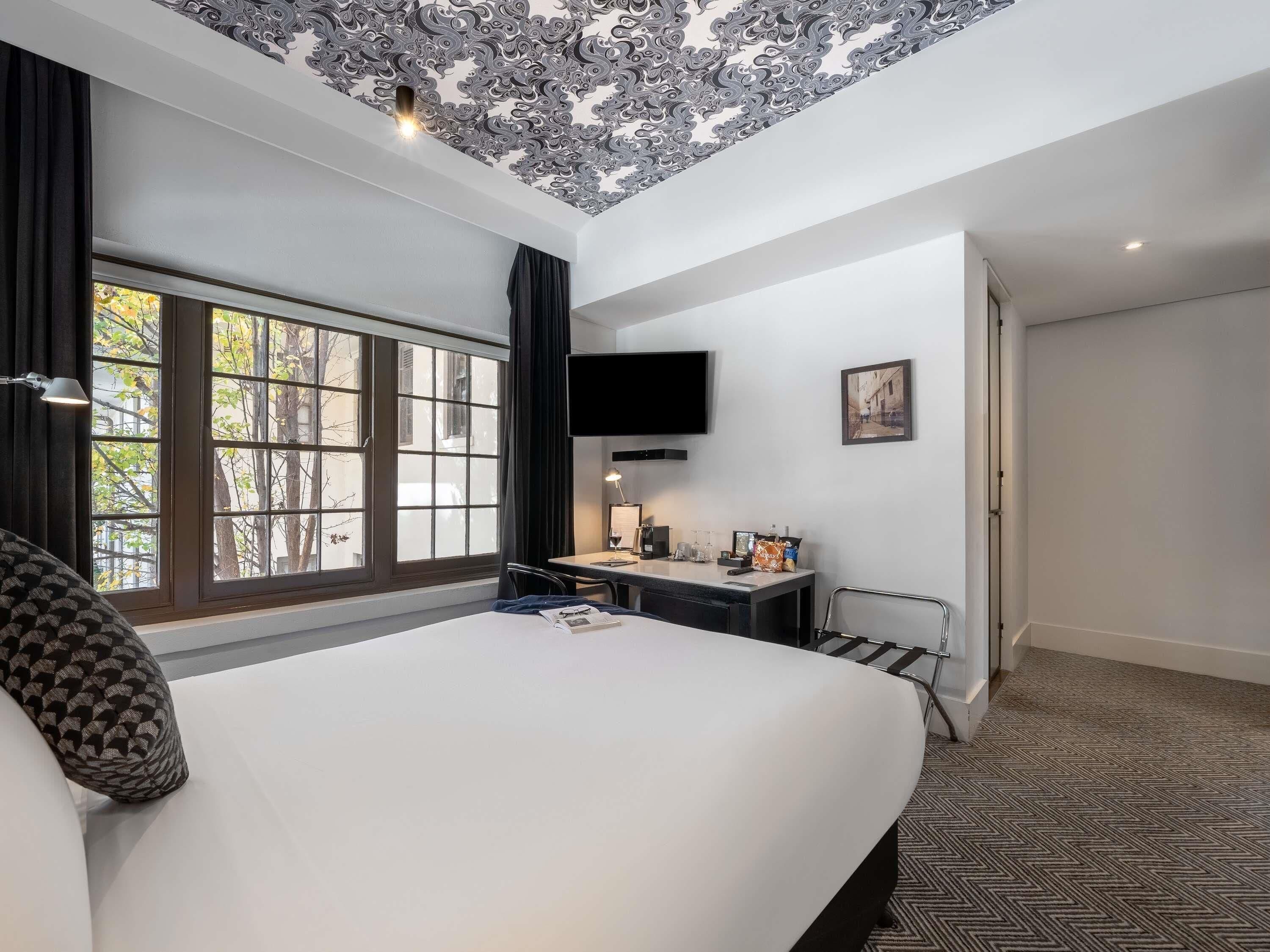 A hotel room with a large bed, a desk with a chair, a TV mounted on the wall, and a window with curtains.