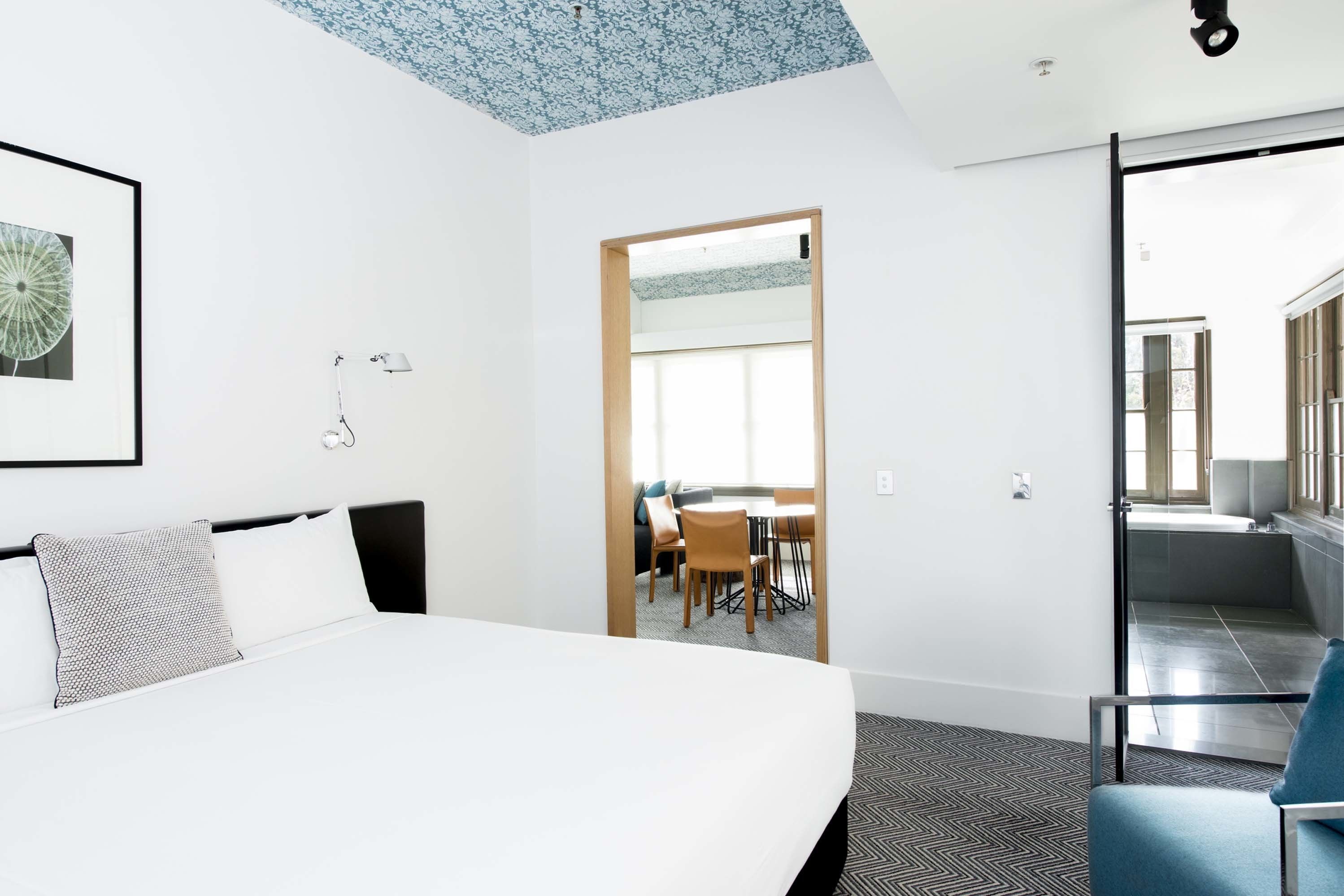 Ein Hotelzimmer mit einem großen Bett, einem Schreibtisch mit Stühlen, einem Badezimmer mit Spiegel und Waschbecken sowie Blick auf den Flur.