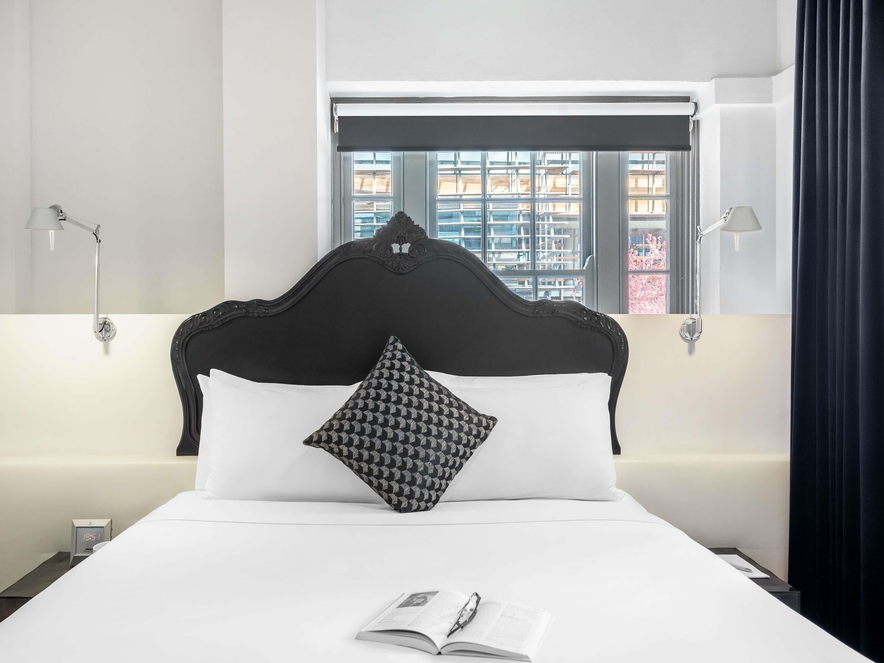 Una cama bien hecha con un cojín estampado, un libro y una pluma encima, una ventana con vistas a edificios, y dos lámparas fijadas a la pared.
