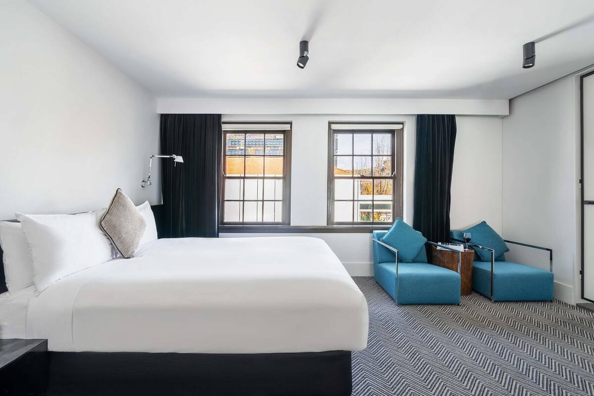 A hotel room with a large bed, two windows, a desk, a chair, and a sofa.