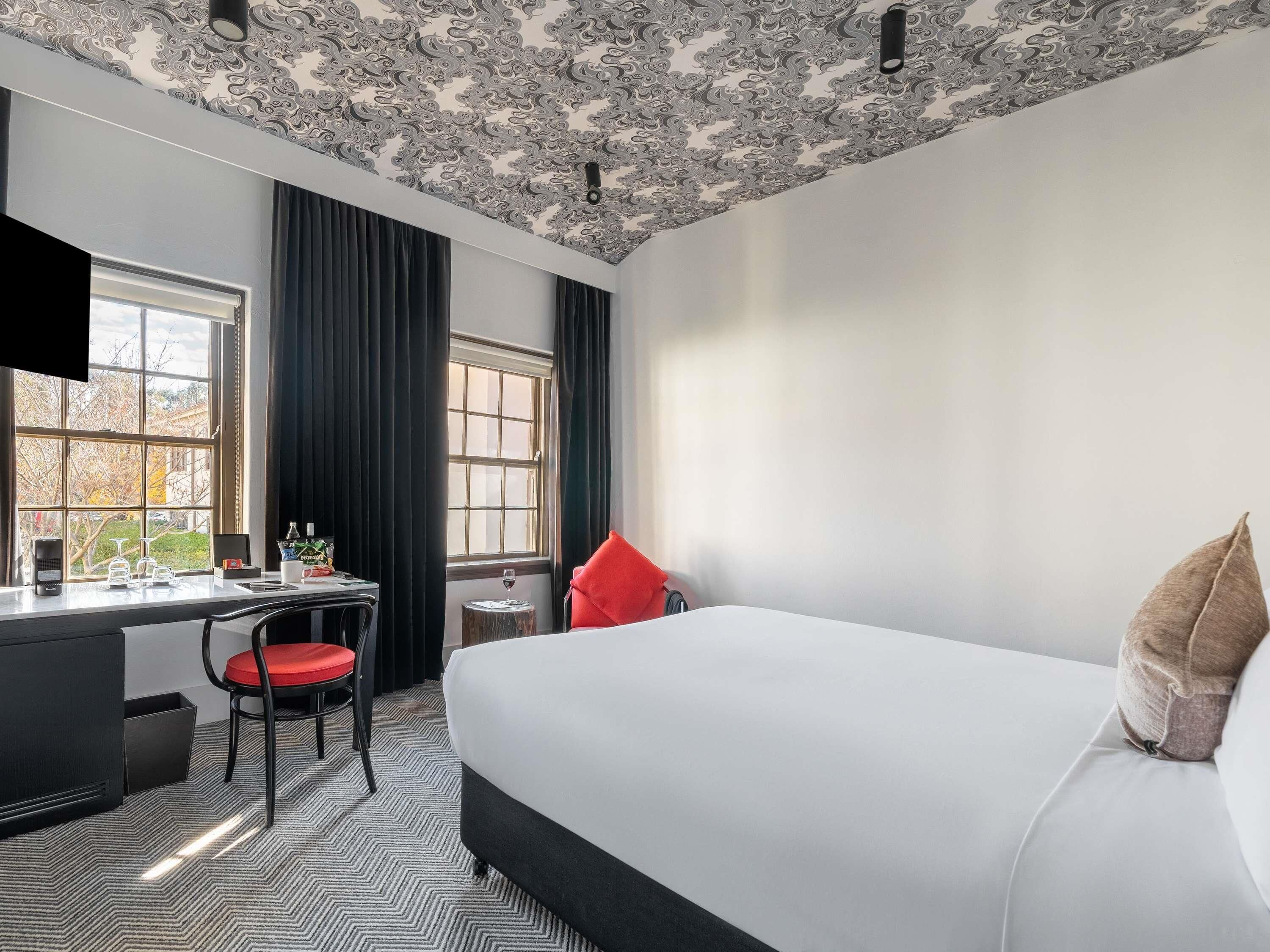 A hotel room with a large bed, a desk, a chair, a small table with a wine glass, and a window with curtains.