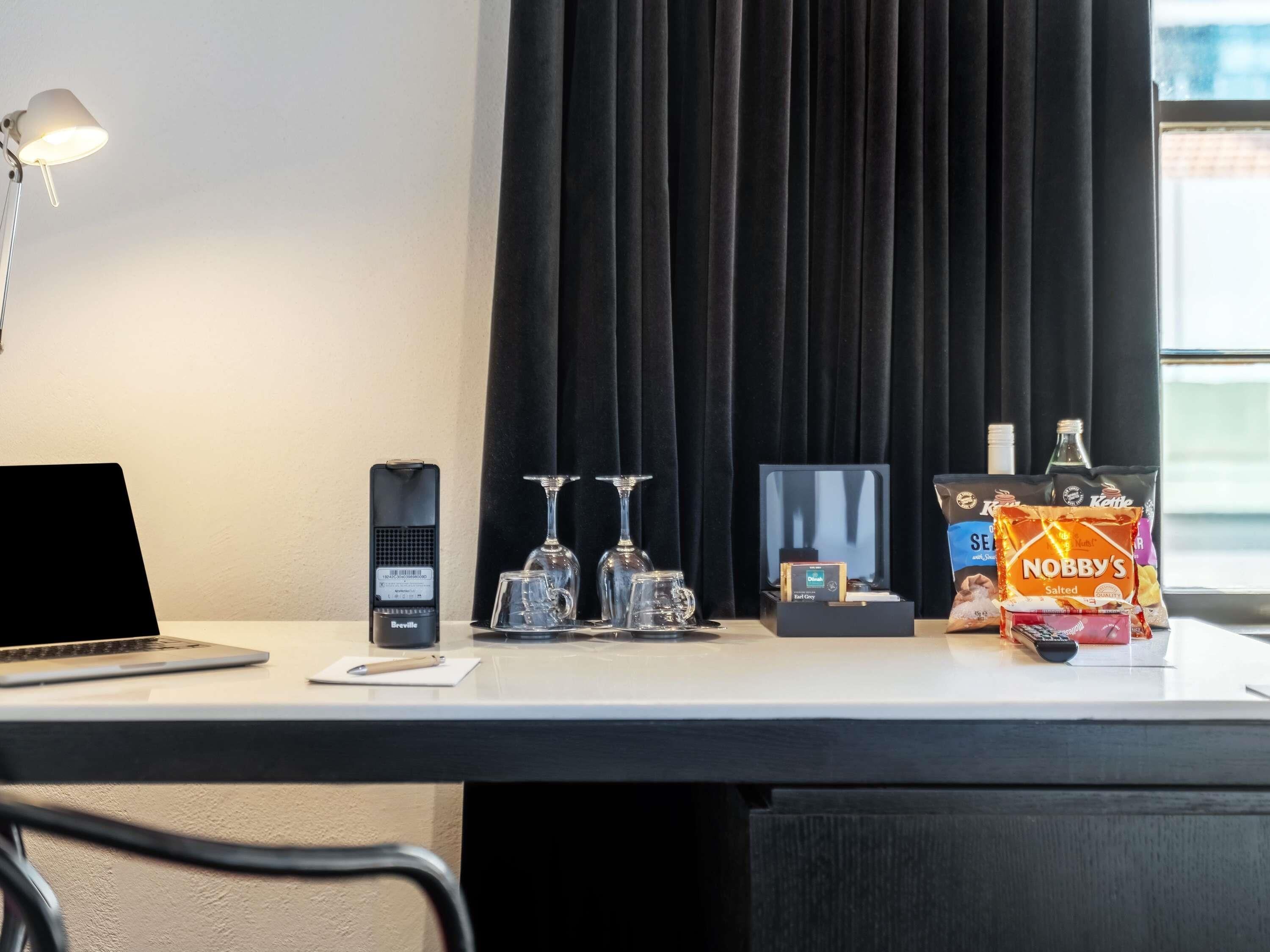 A hotel room desk with a laptop, a Breville coffee maker, two wine glasses, a box of Nobby's Salted snacks, and a bottle of liquor.