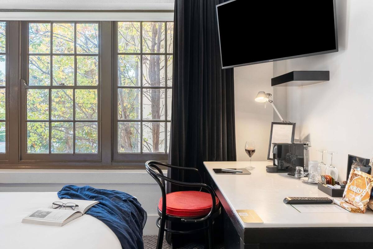 A hotel room with a bed, a desk with a computer, a chair, a TV mounted on the wall, and a window with a view of trees.