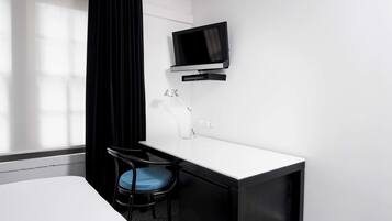 A hotel room with a white desk, a black chair, a TV mounted on the wall, and a window with curtains.