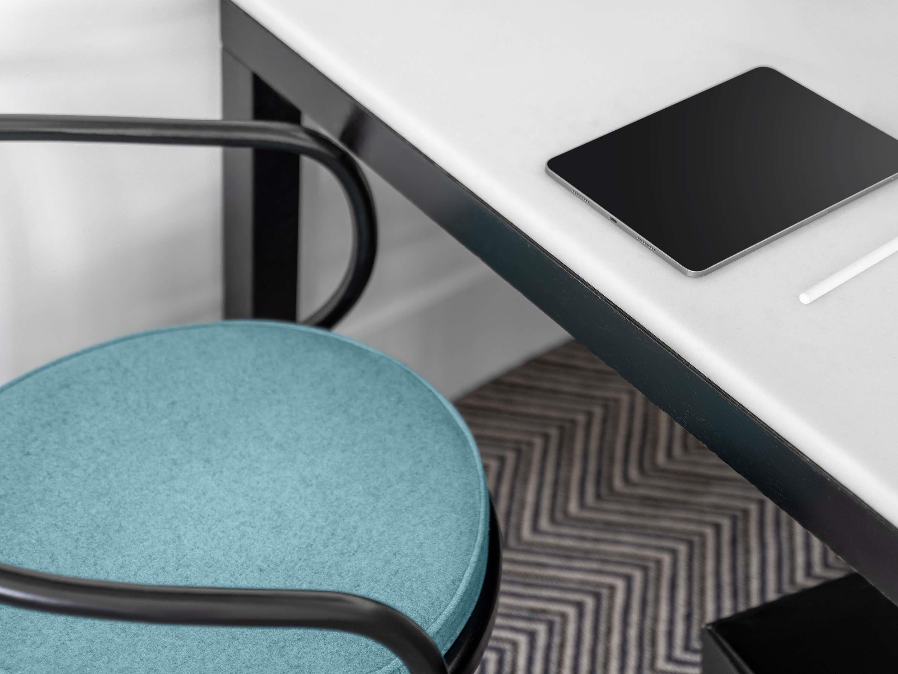 Ein moderner Bürostuhl mit türkisfarbenem Sitzkissen, ein schwarzes Tablet auf einem weißen Schreibtisch und ein strukturierter Boden.