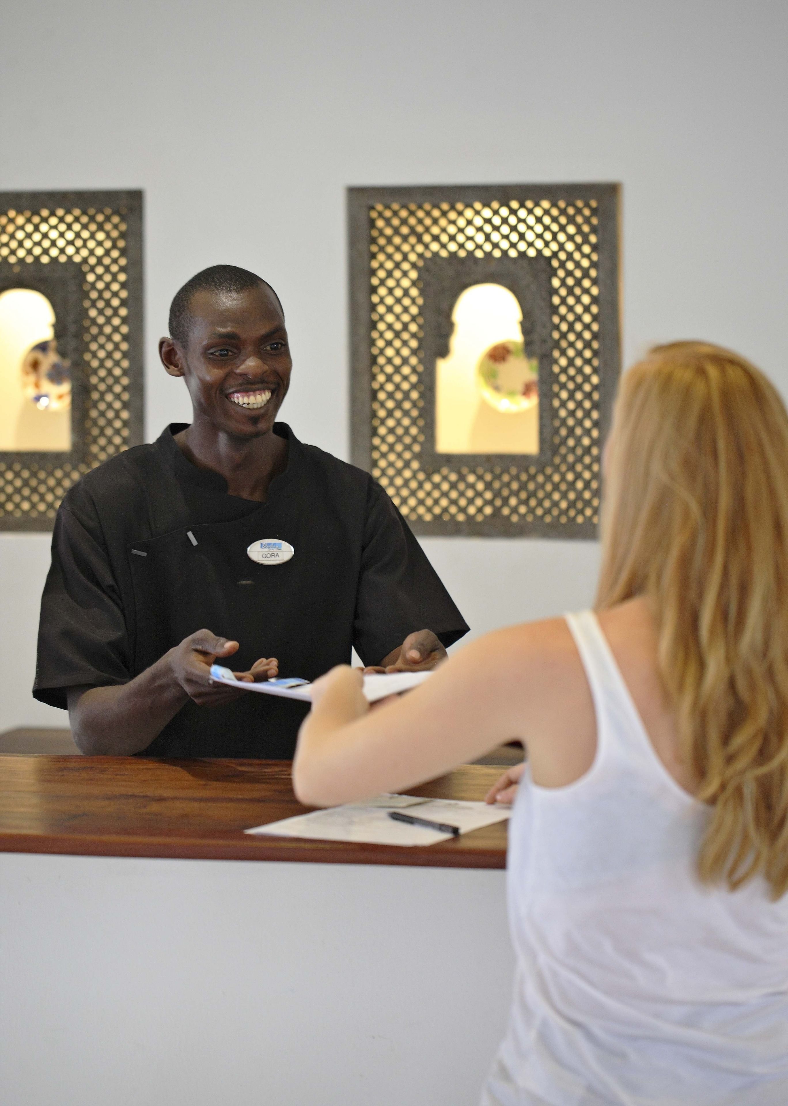 A reception desk with a person in a black uniform and a woman in a white top. There are framed pictures on the wall behind the desk.
