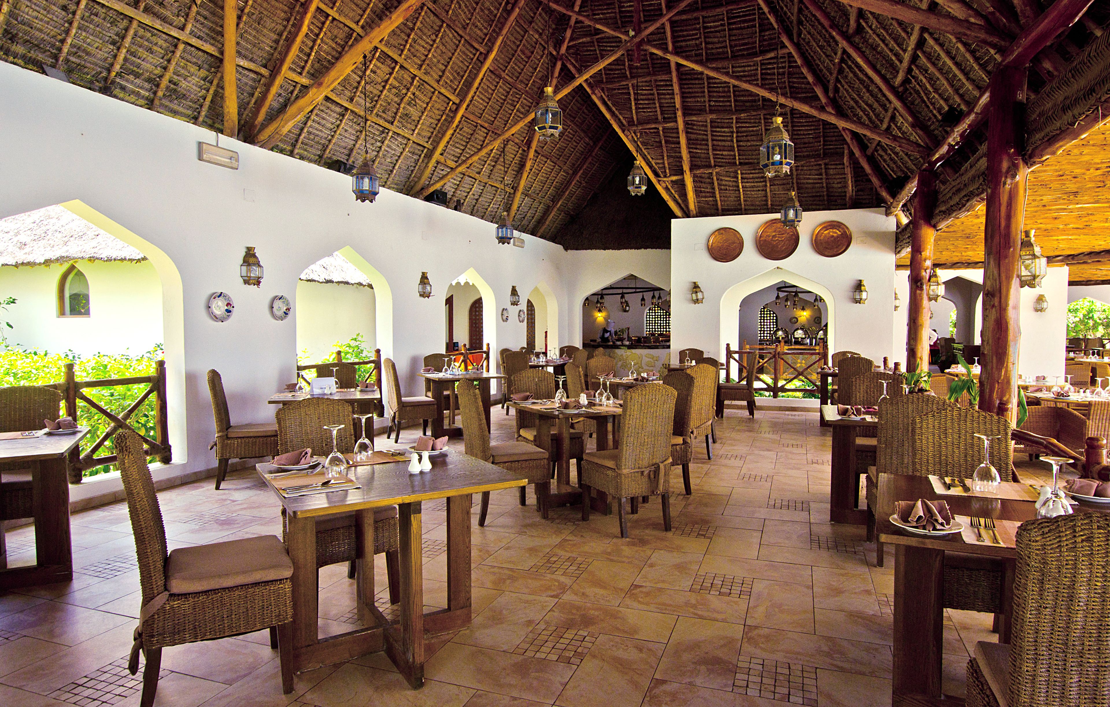 An indoor dining area with wooden tables and wicker chairs, a thatched roof, and decorative lanterns.