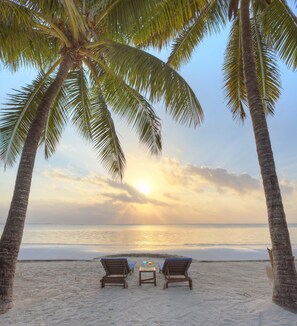 Une plage avec deux chaises longues en bois et une petite table, entourée de palmiers et offrant une vue sur l’océan.