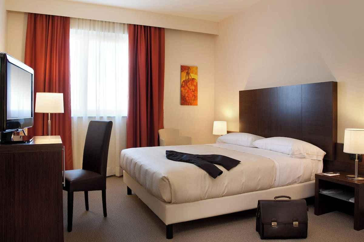 Hotellihuone, jossa on sänky, työpöytä tietokoneella, tuoli, yöpöytä lampun kanssa ja taulu seinällä.