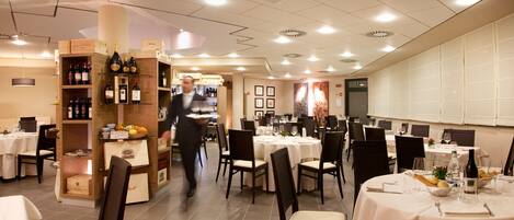 Un restaurant avec de nombreuses tables dressées pour le service, un présentoir à vins et un serveur qui s’affaire.
