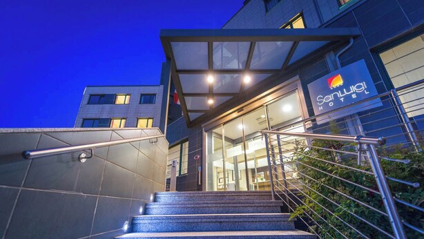 A hotel entrance with a glass facade, metal railings, and a staircase leading up to the entrance.