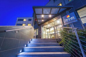A hotel entrance with a glass facade, metal railings, and a staircase leading up to the entrance. - Hotel San Luigi (Beinasco)