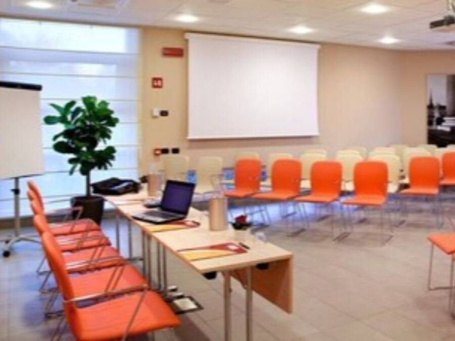 A conference room with rows of orange and beige chairs, a projector screen, and a laptop on a table.