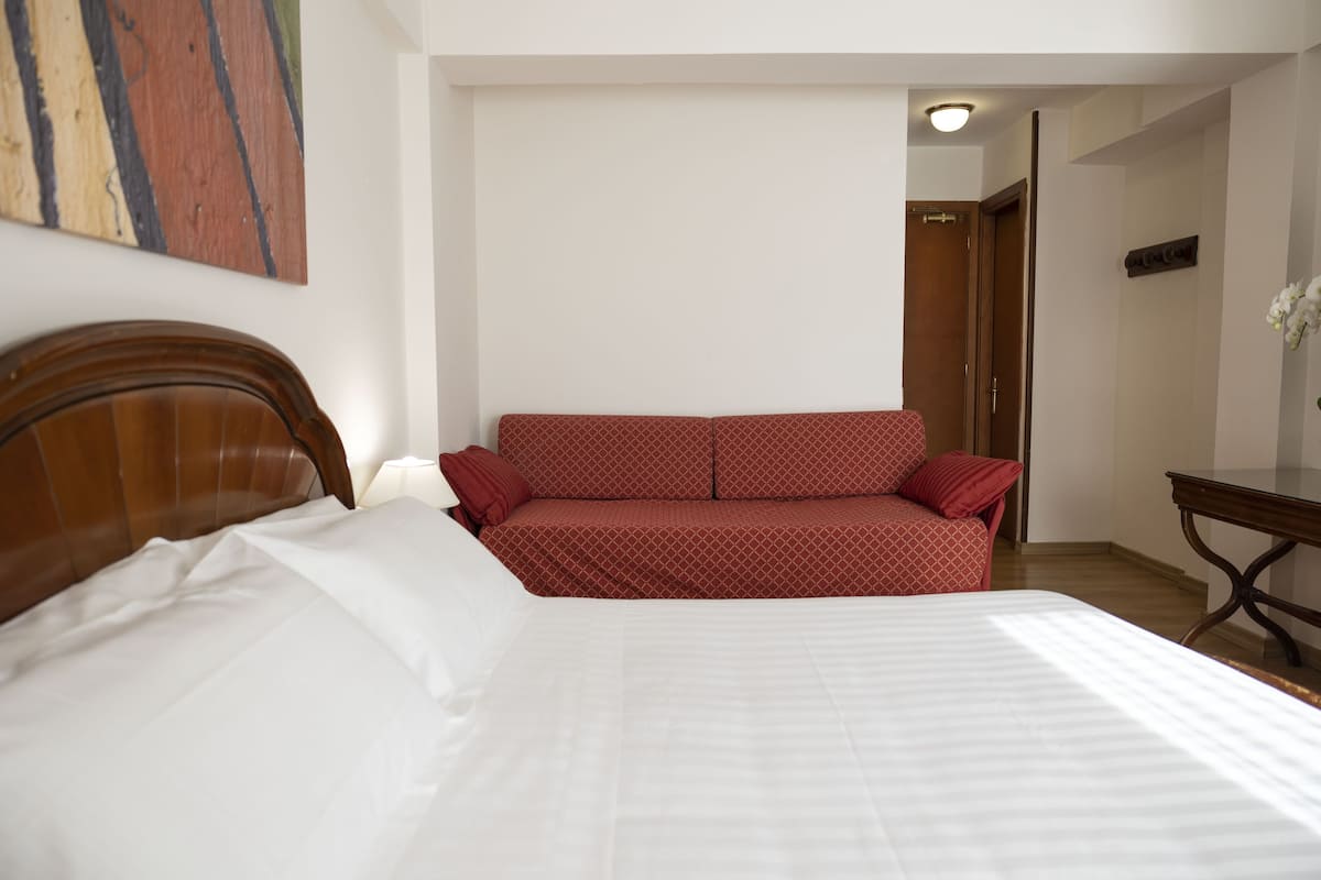 A bedroom with a bed, a red patterned sofa, a wooden headboard, a nightstand, and a wall-mounted artwork.