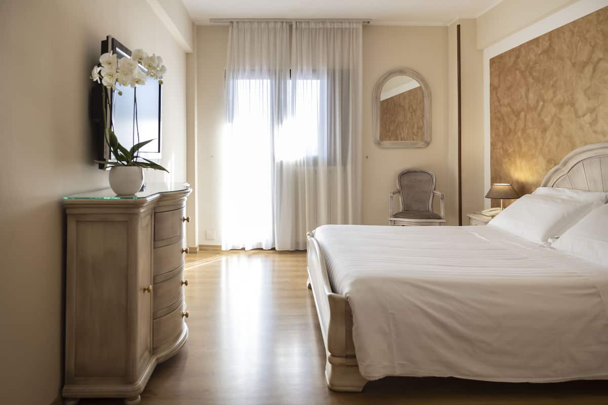 A bedroom with a large bed, a dresser, a chair, a mirror, and a vase of flowers.