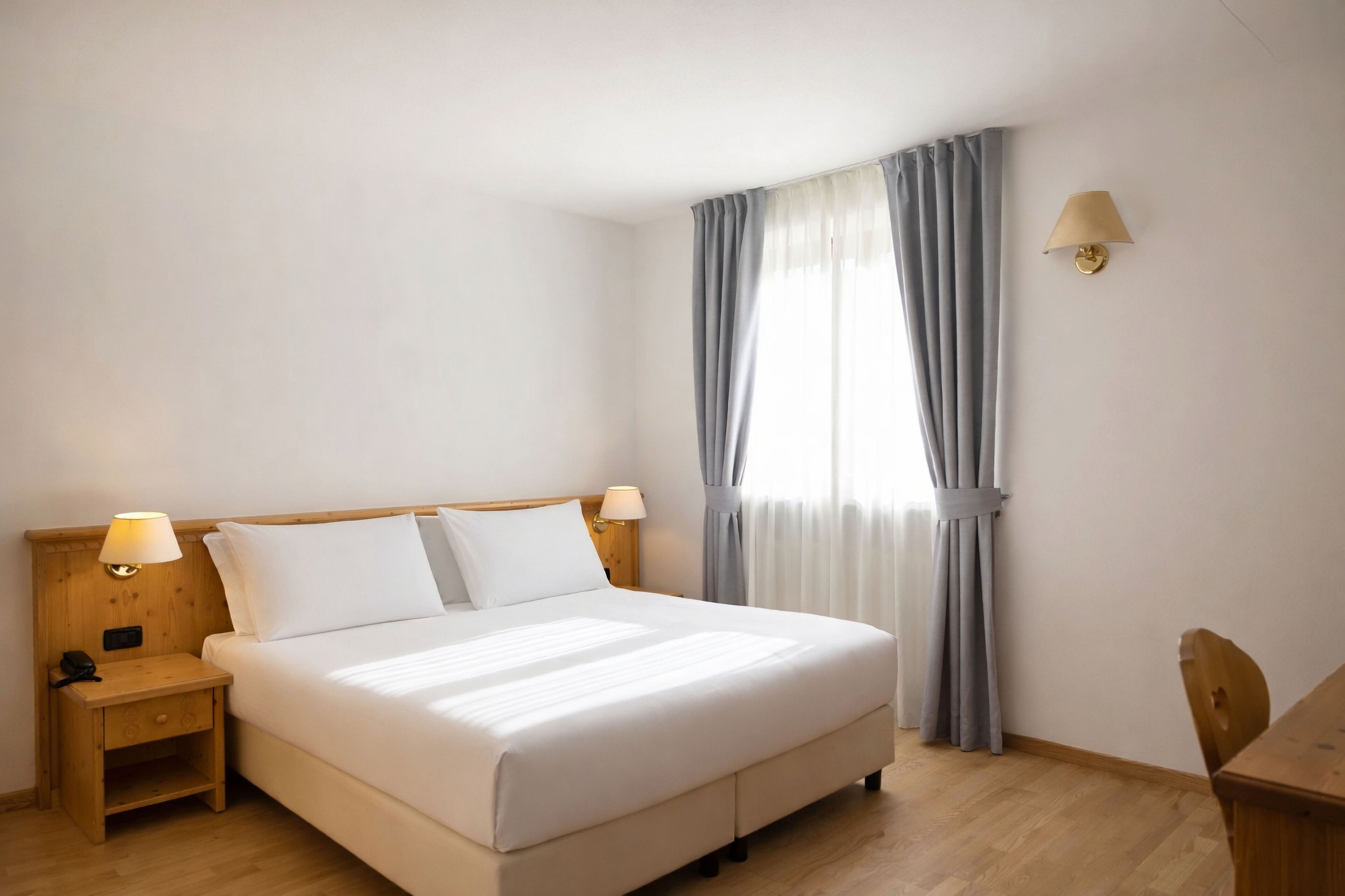 A hotel room with a single bed, a nightstand with a lamp, a wooden chair, and a window with curtains.