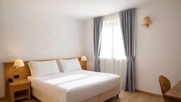 Ein Hotelzimmer mit einem Einzelbett, einem Nachttisch mit Lampe, einem Holzstuhl und einem Fenster mit Vorhängen.