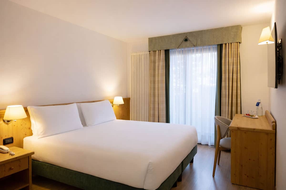A hotel room with a large bed, two bedside lamps, a wooden desk, a chair, and a TV on the wall.