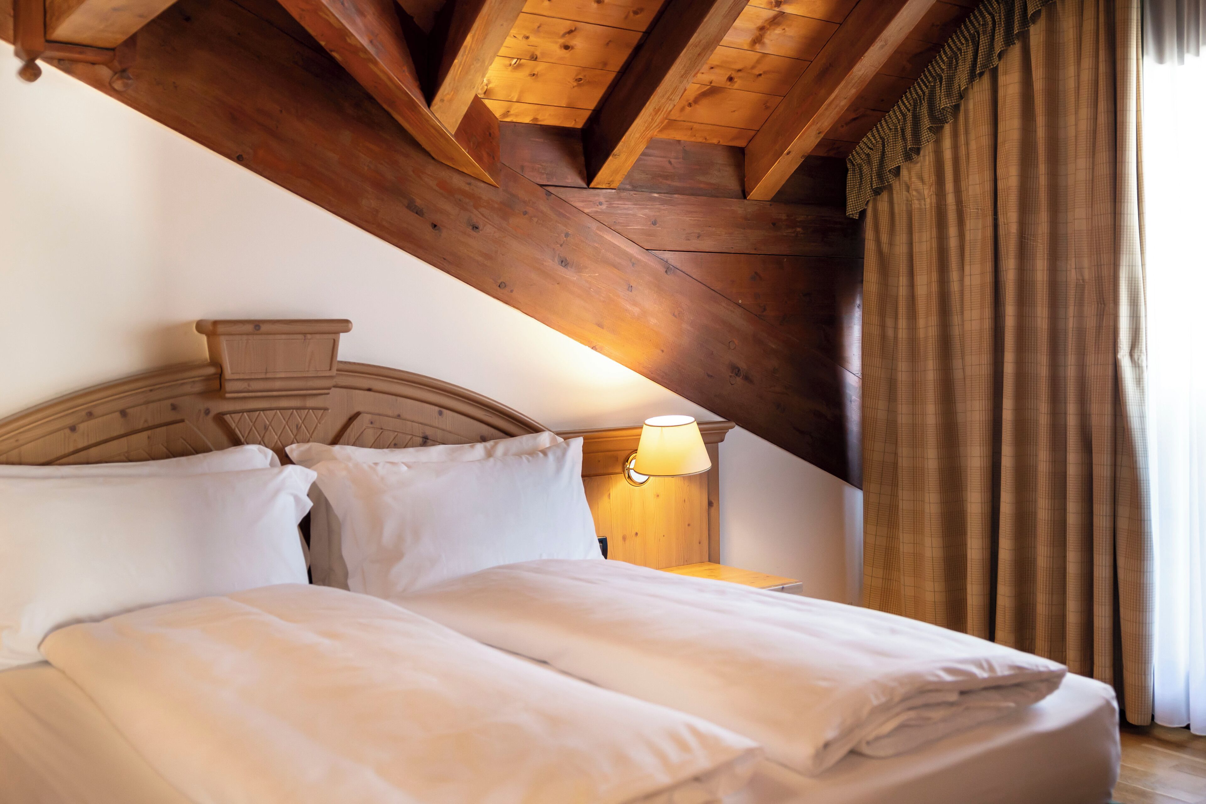 A bedroom with a wooden ceiling, a bed with white bedding, a nightstand with a lamp, and a window with curtains.