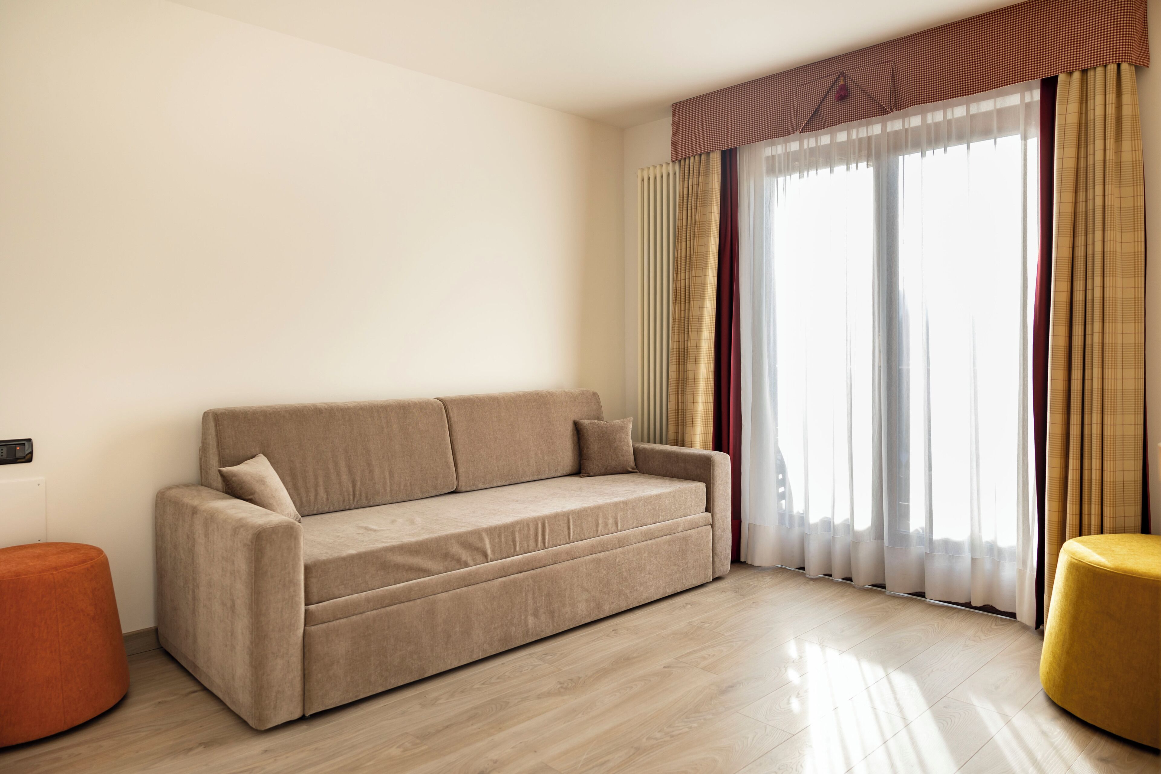 A living room with a beige sofa, a round orange ottoman, and a large window with sheer curtains.