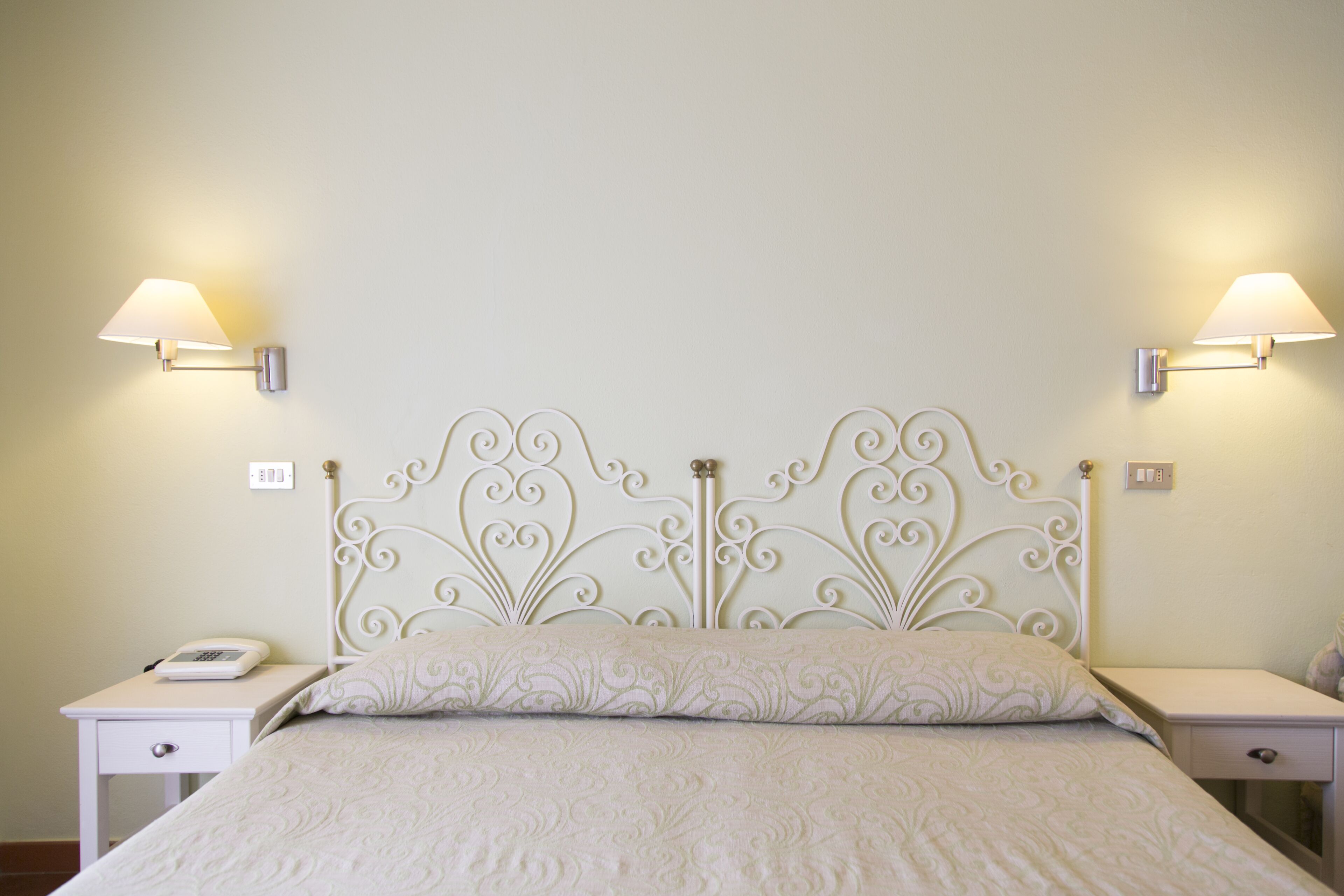 Una cama bien tendida con una colcha estampada, un cabecero de metal blanco con diseños intrincados, dos mesitas de noche con lámparas, y una mesita de noche pequeña con un teléfono.